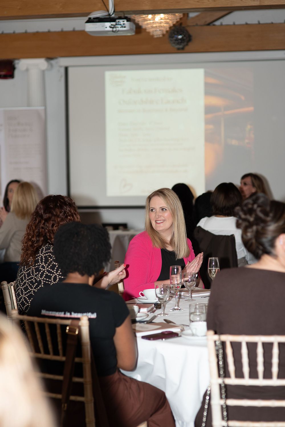 Women networking at a professional luncheon event, with a smiling woman in pink blazer seated at a white-clothed table.