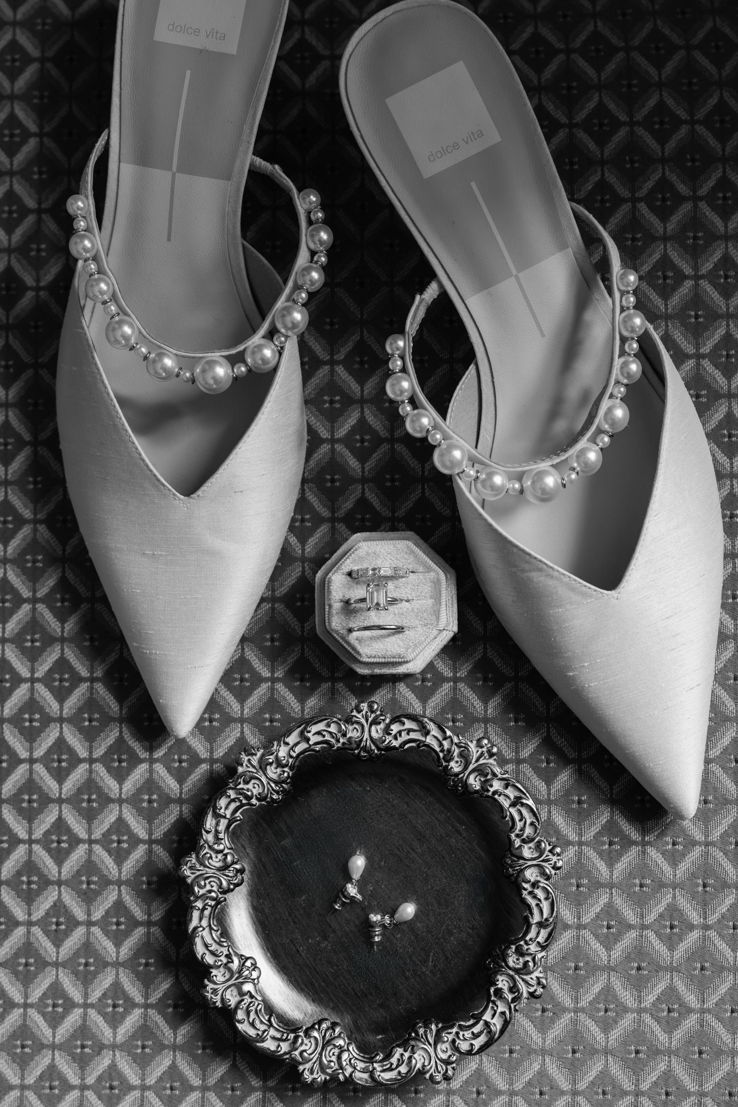 Black and white photo of pearl-adorned bridal heels with an engagement ring and octagonal ring box on patterned fabric.