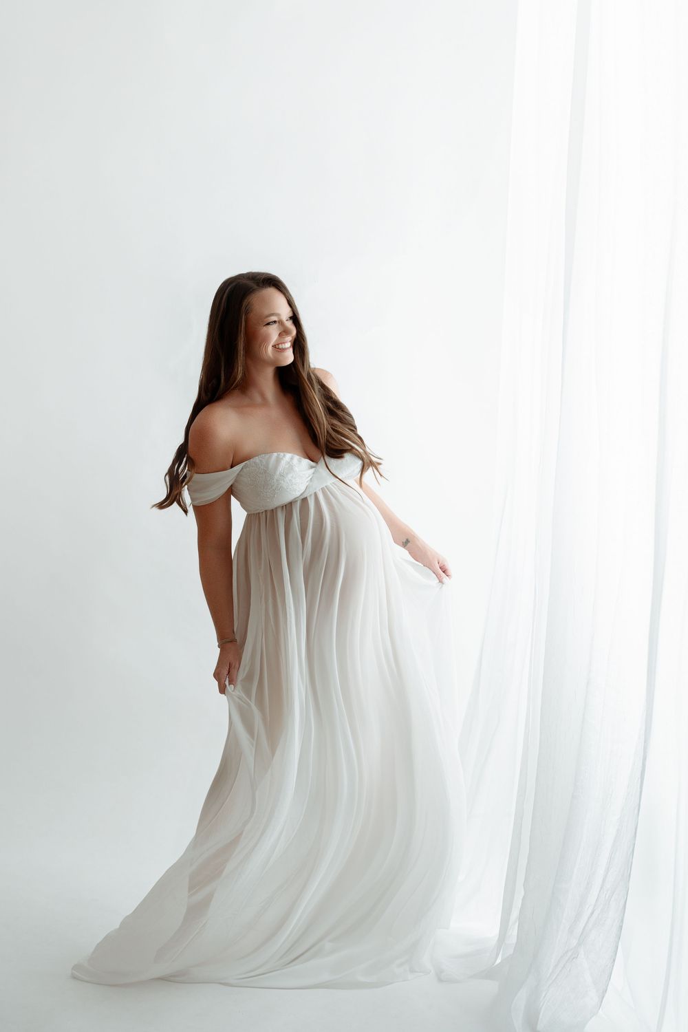 Pregnant woman in flowing white off-shoulder gown poses gracefully near sheer curtains in bright studio.