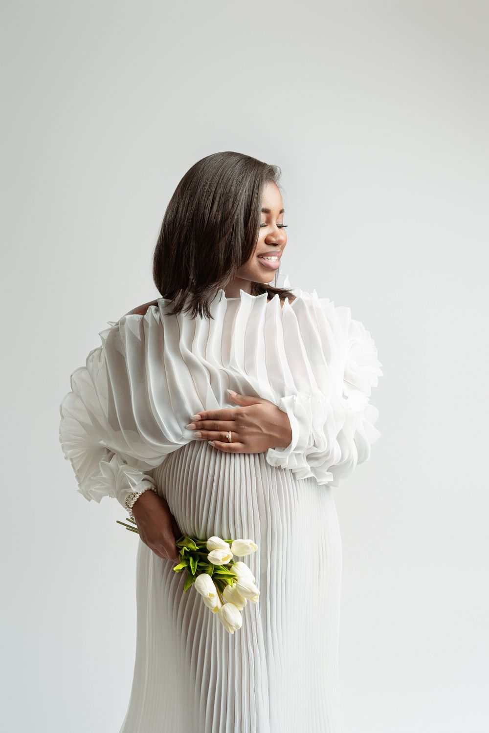 Smiling pregnant woman in white pleated dress holding white roses against light background.