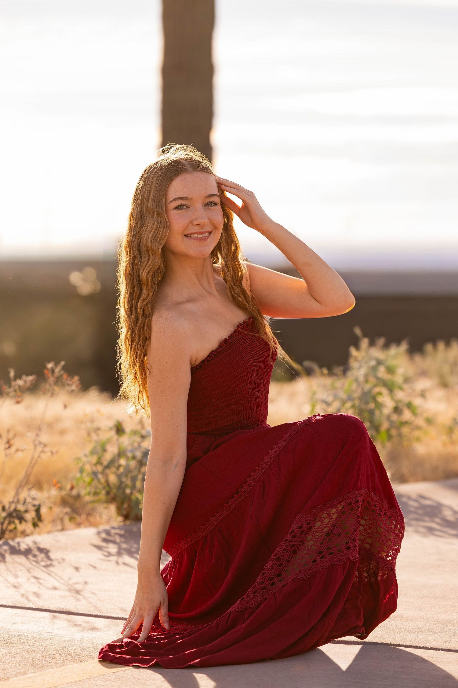 Young woman in red maxi dress sitting outdoors in golden sunset light, smiling with hand in hair.