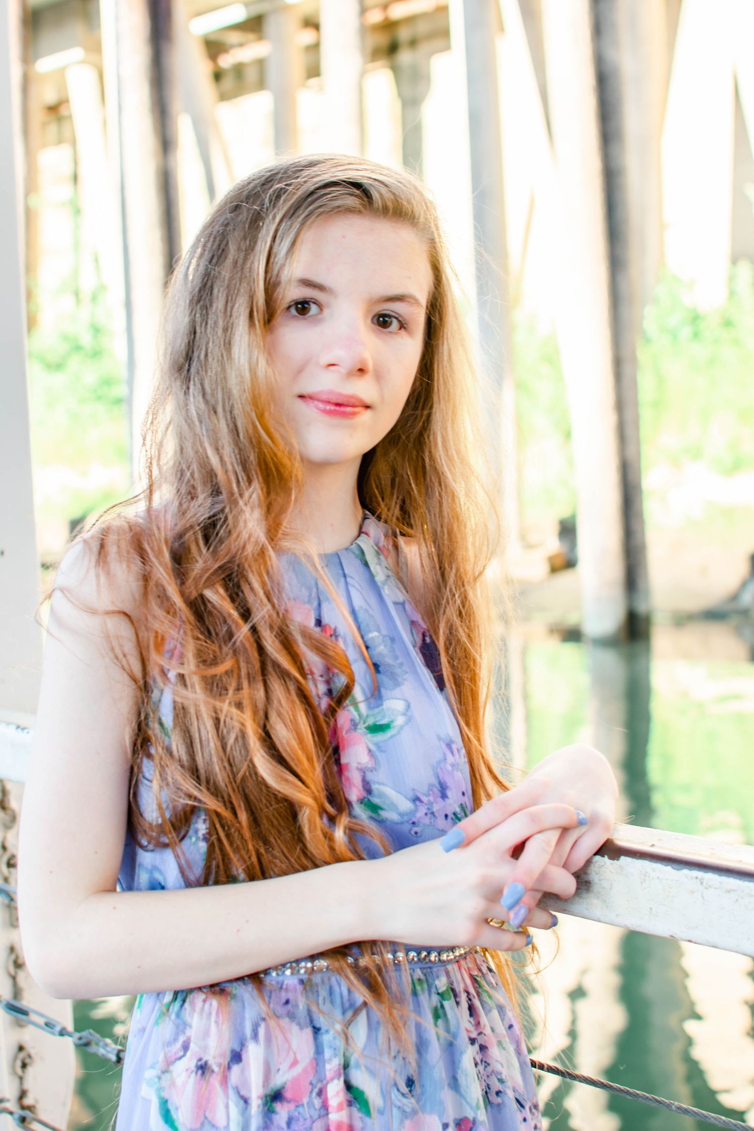 Young woman with long auburn hair in floral dress poses by white railing near waterfront pier.