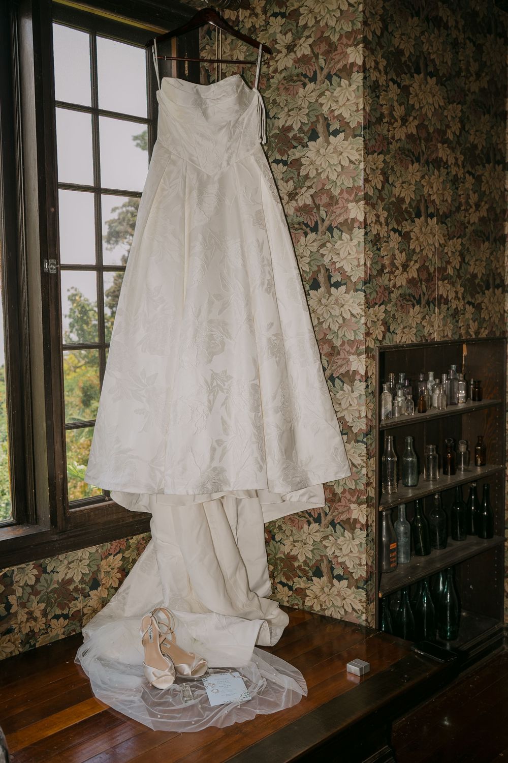 White strapless wedding dress hanging by window with bridal heels and veil displayed on wooden table.