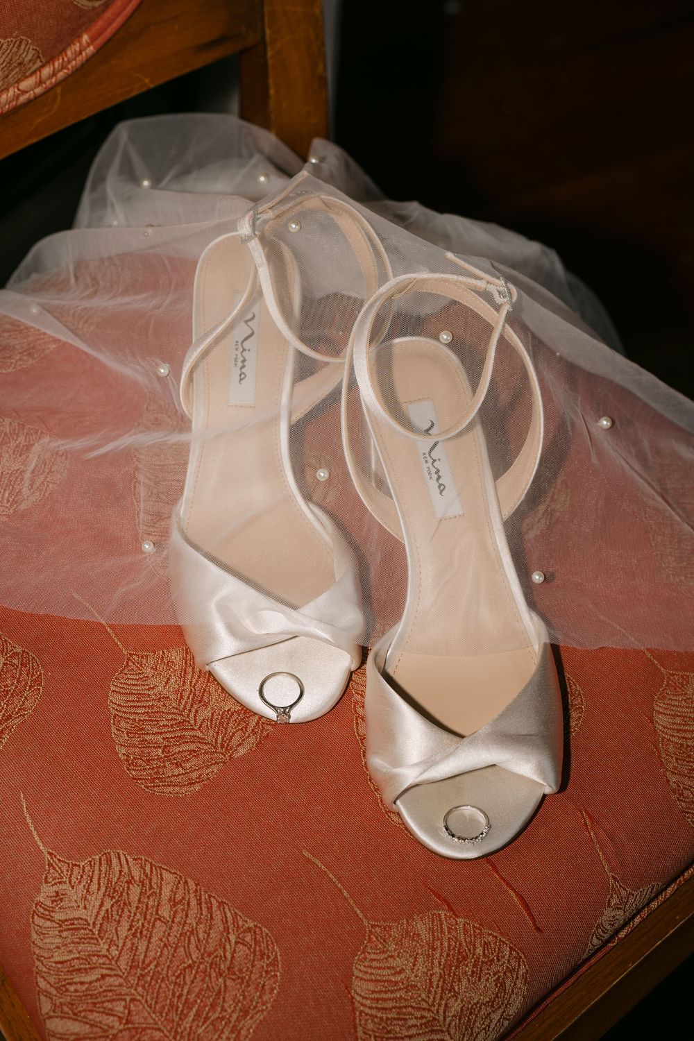 Elegant white satin bridal heels with ankle straps and pearl details resting on a floral chair with a wedding veil.