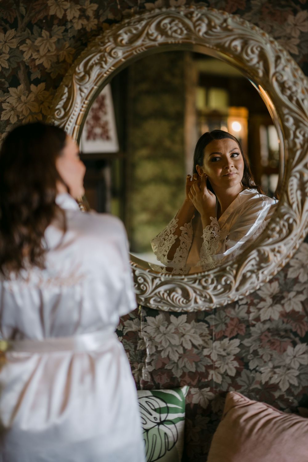 Bride in white lace robe adjusting earrings while looking into ornate vintage round mirror in floral wallpapered room.