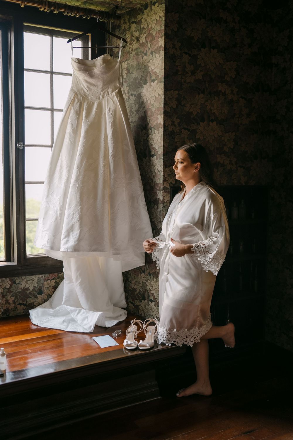 Bride in white lace robe admiring her wedding gown hanging by a window with heels below.