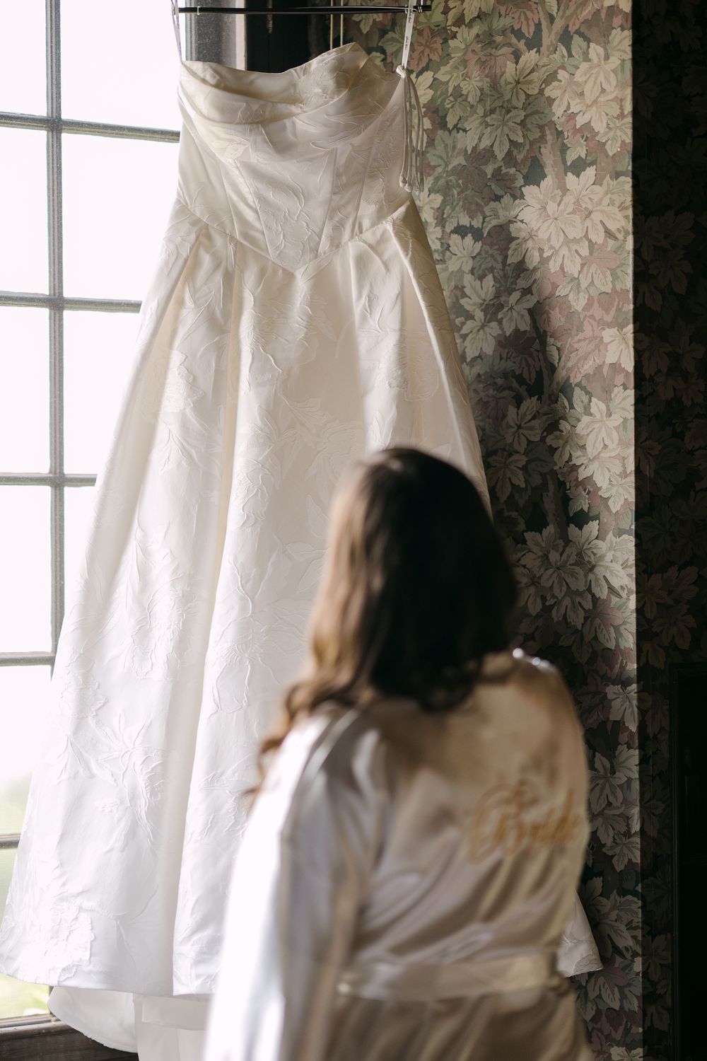Bride in white robe gazing at elegant strapless wedding gown hanging on floral wallpaper near window.
