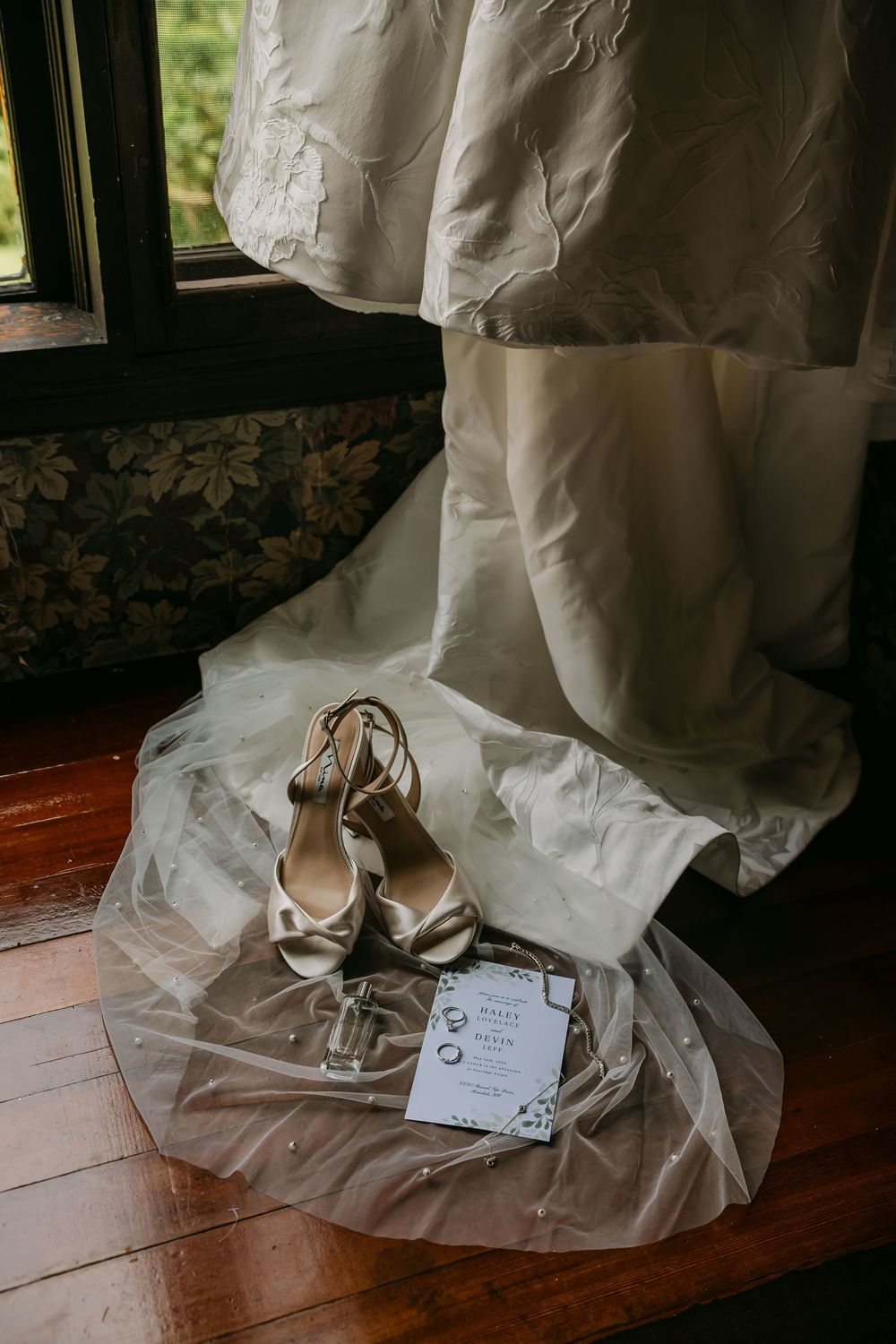 Bridal wedding dress hanging near window with elegant heels and invitation on tulle veil on wooden floor.
