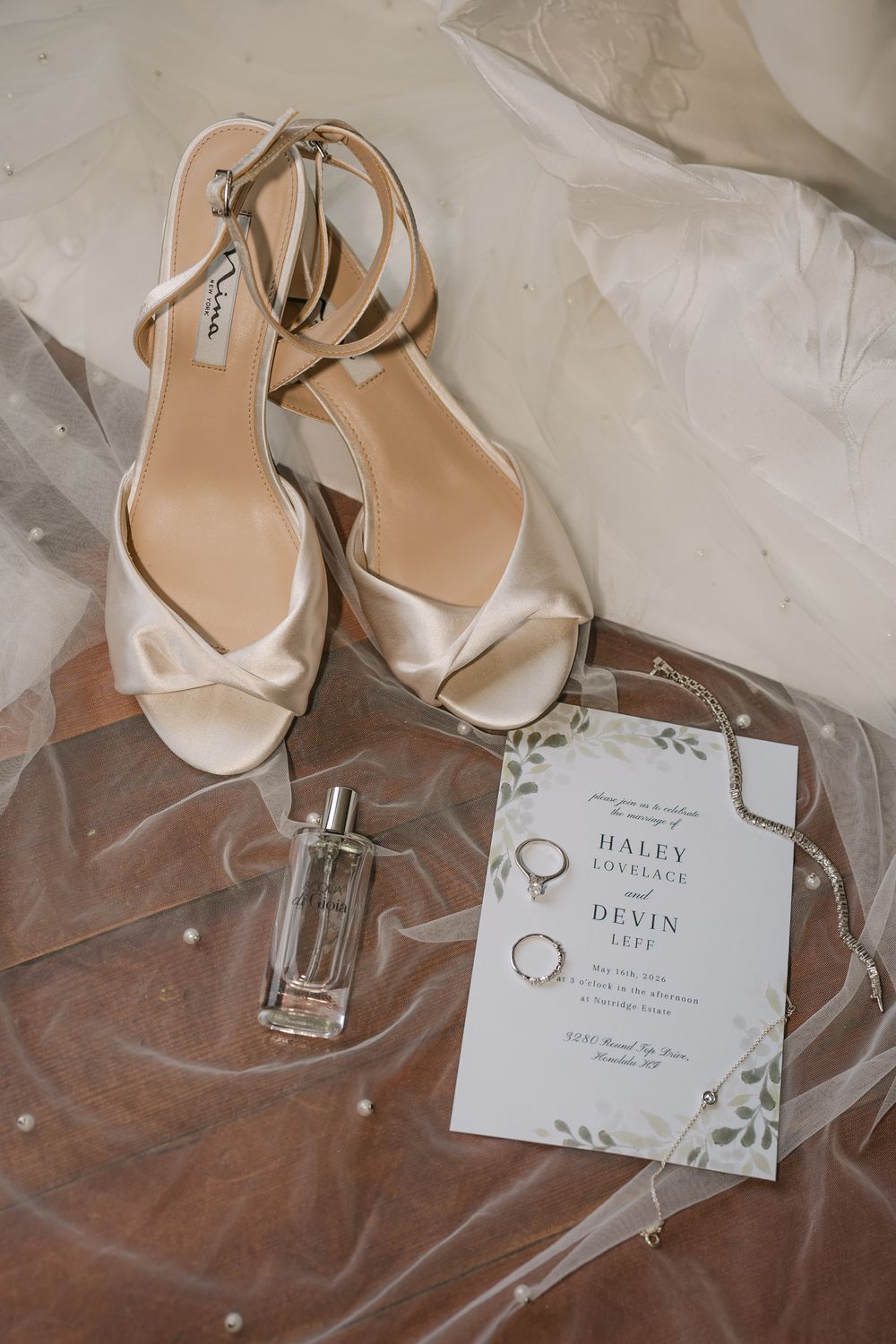 Elegant bridal flat lay with champagne heels, perfume, rings, and a floral wedding invitation on sheer veil fabric.