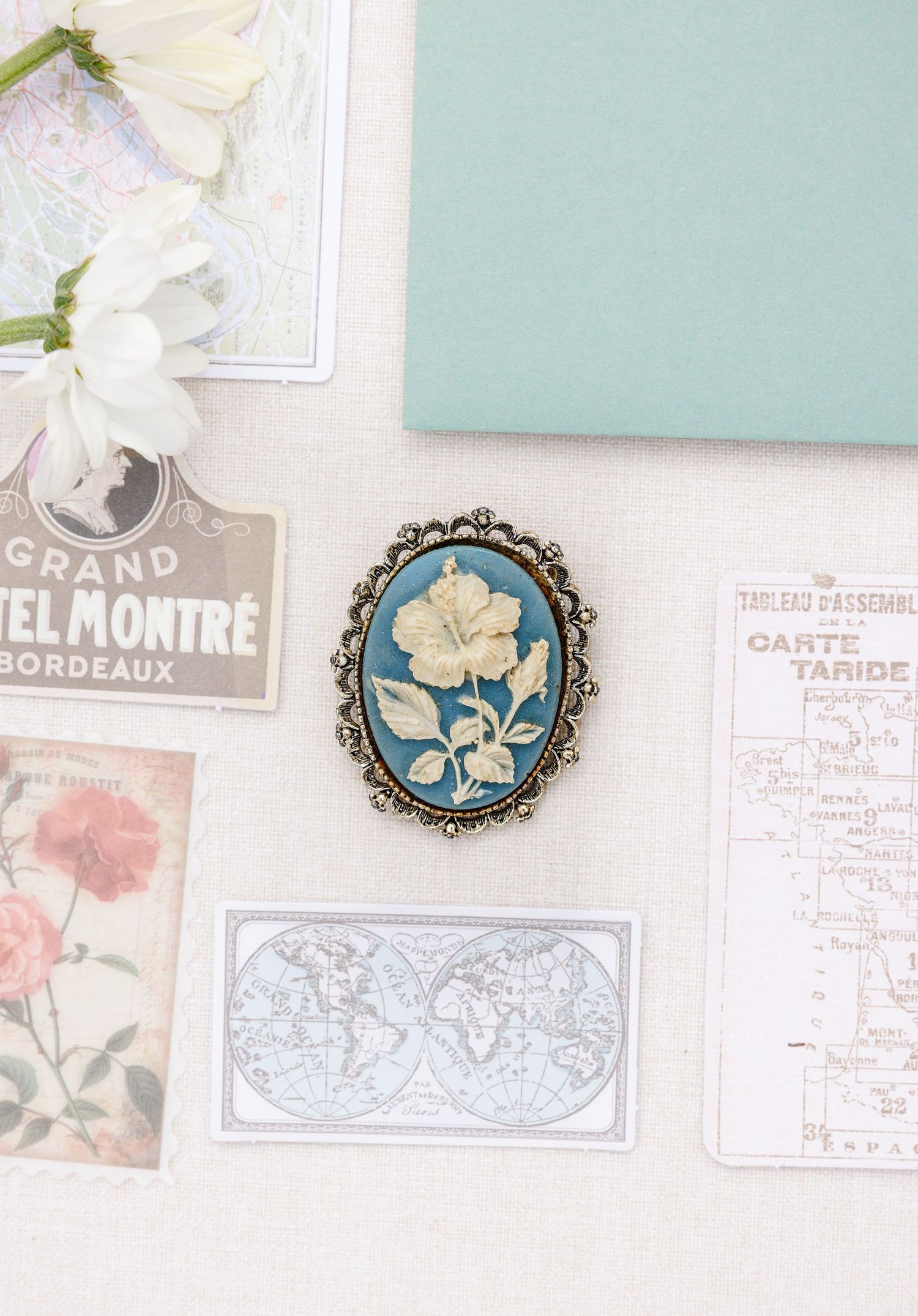 Vintage blue cameo brooch with ivory floral design in ornate gold setting on flatlay with postcards and flowers.
