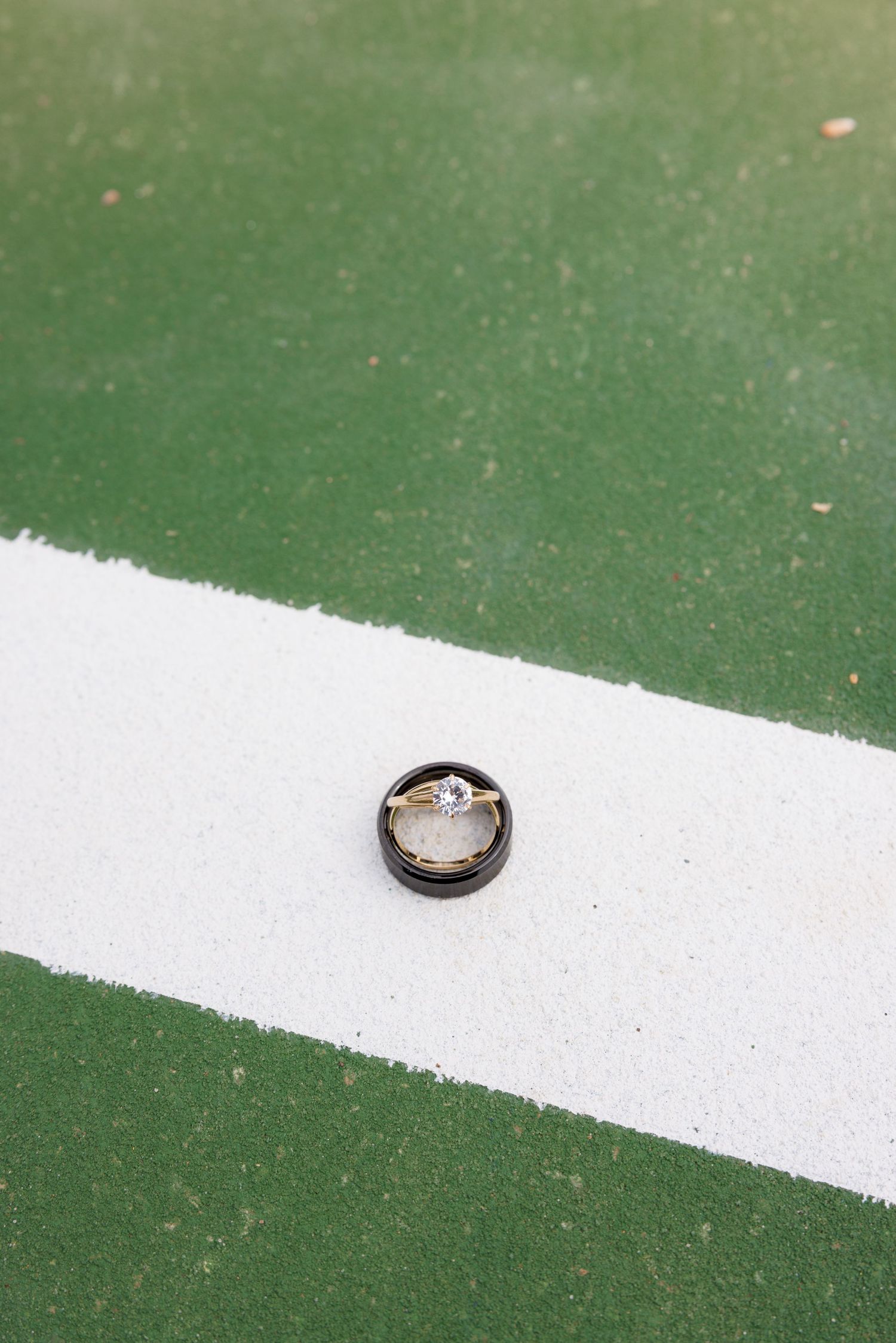 Wedding rings, a diamond engagement ring nestled in a black band, placed on a green tennis court white line.