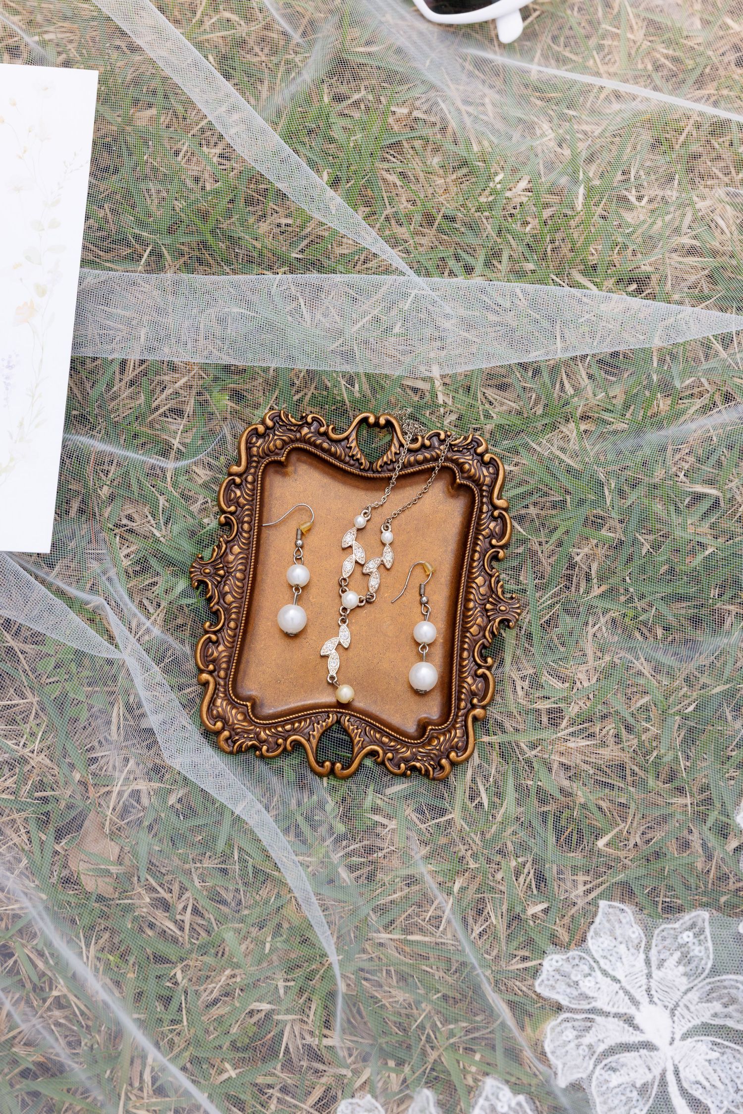 Ornate gold vintage frame displaying pearl drop earrings and delicate floral necklace on grass with sheer bridal veil.