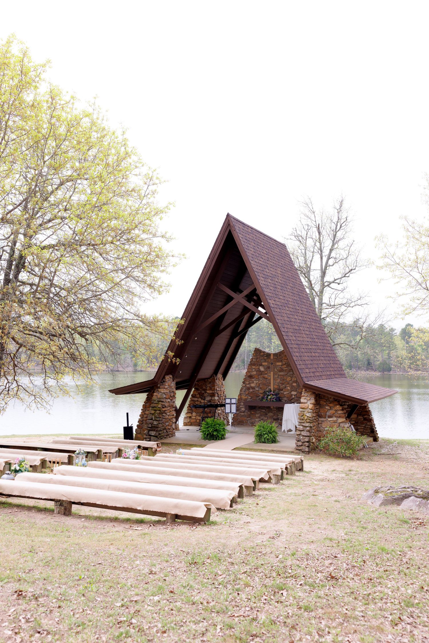Rustic outdoor chapel with A-frame roof beside a serene lake, featuring wooden pew benches and stone walls.