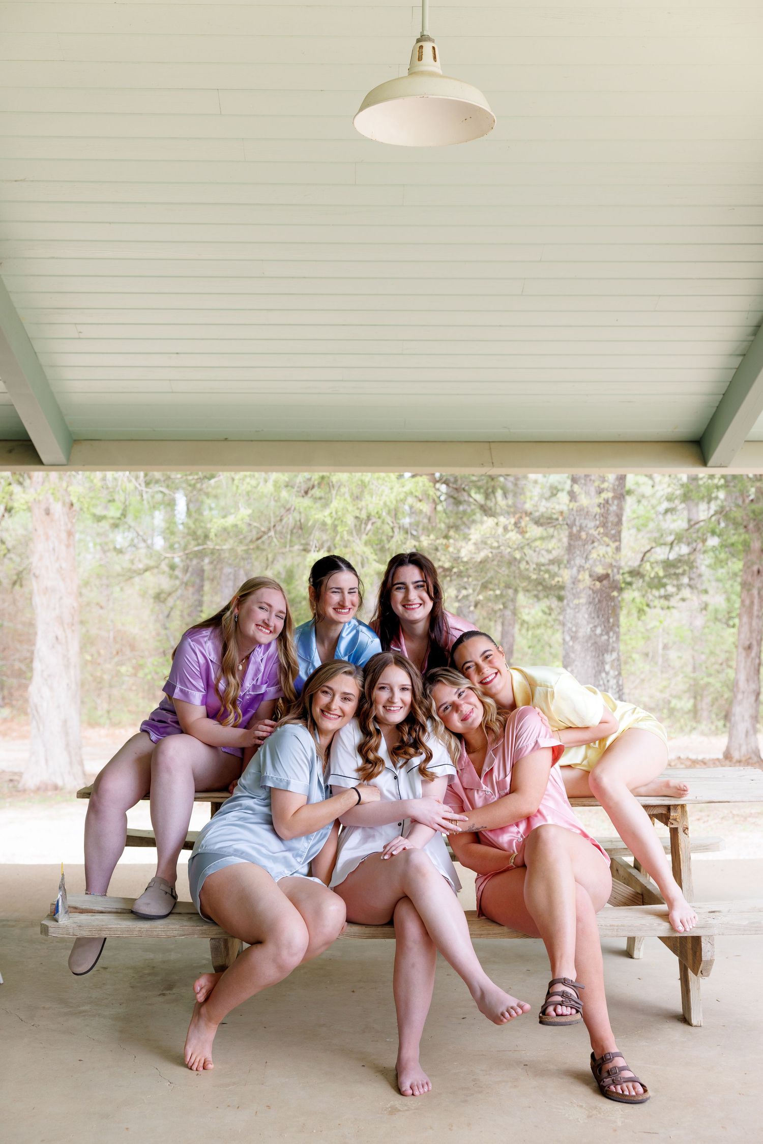 Seven bridesmaids in colorful satin robes smile together on a picnic table under a covered outdoor pavilion.