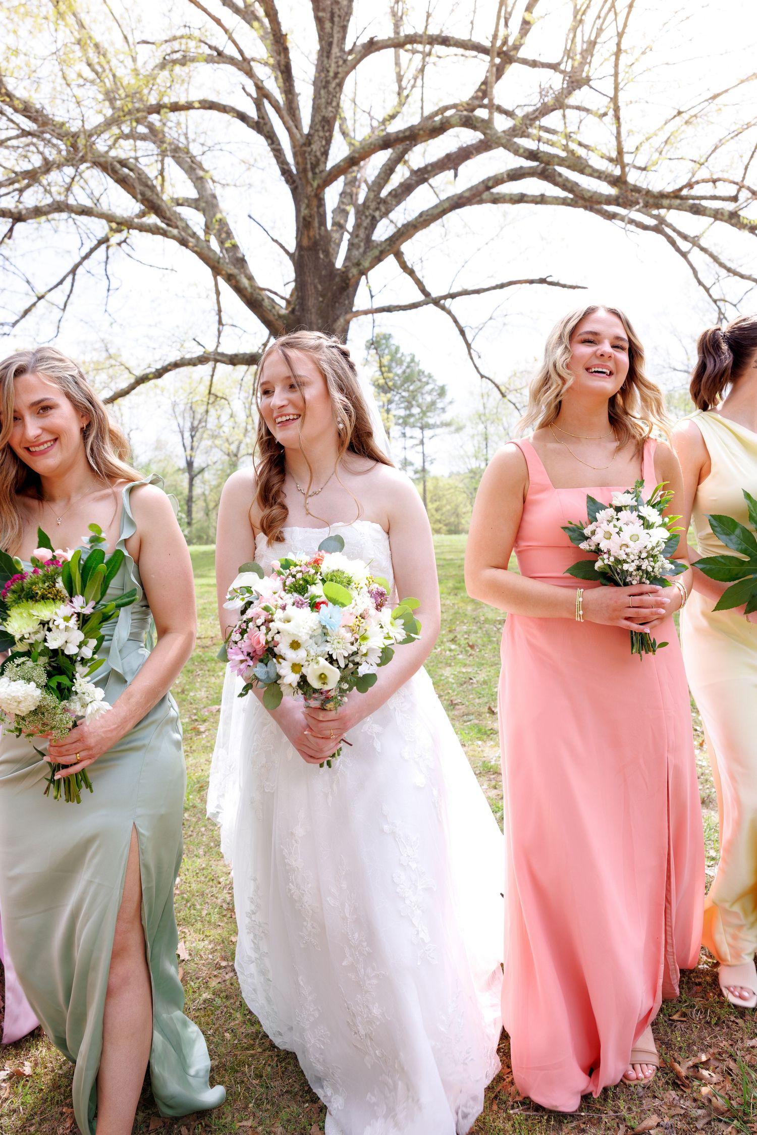 Bride in white gown stands with smiling bridesmaids in sage and pink dresses holding floral bouquets outdoors.