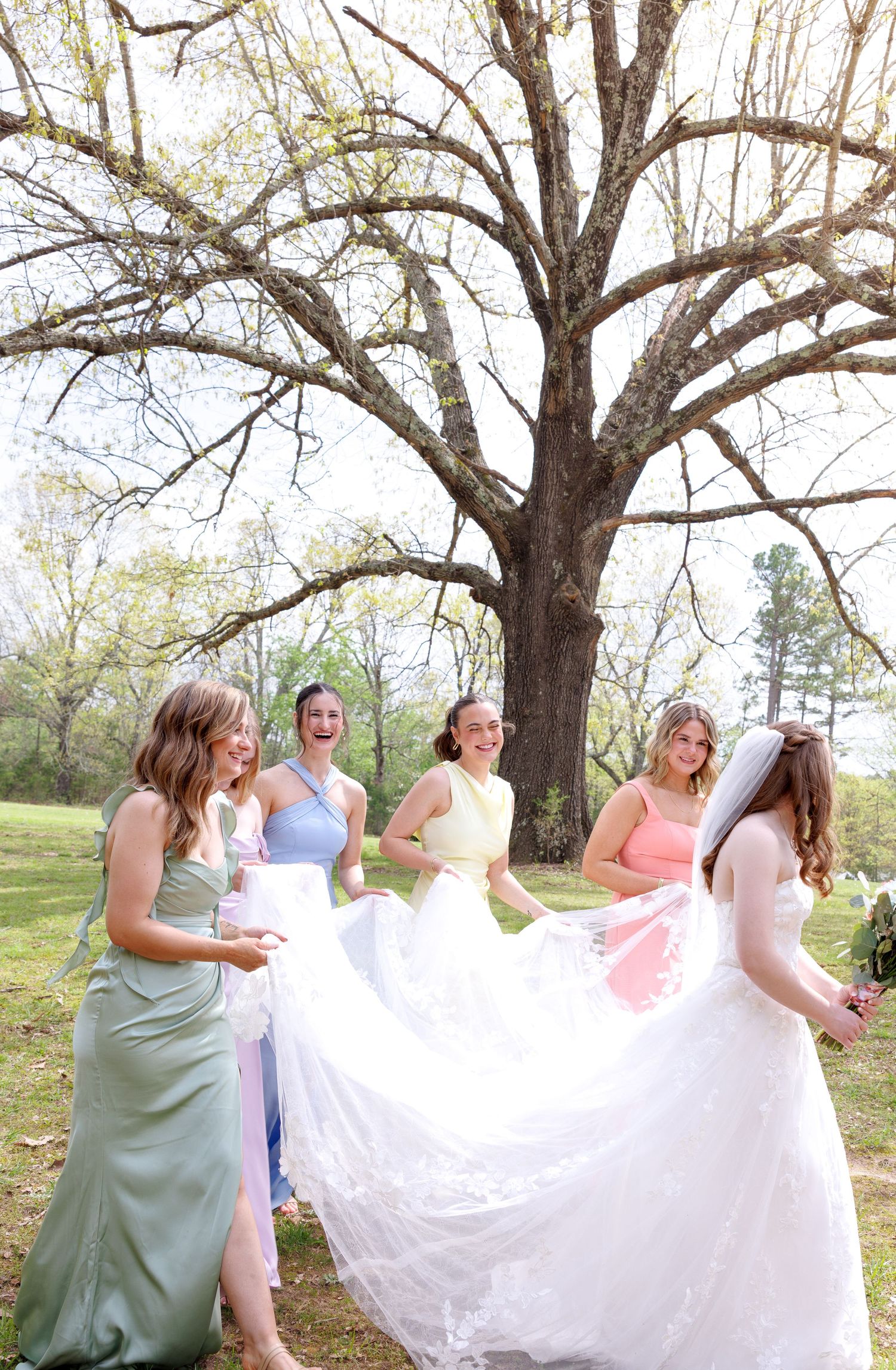Bridesmaids in colorful dresses help a bride with her white gown outdoors near a large tree on a sunny spring day.