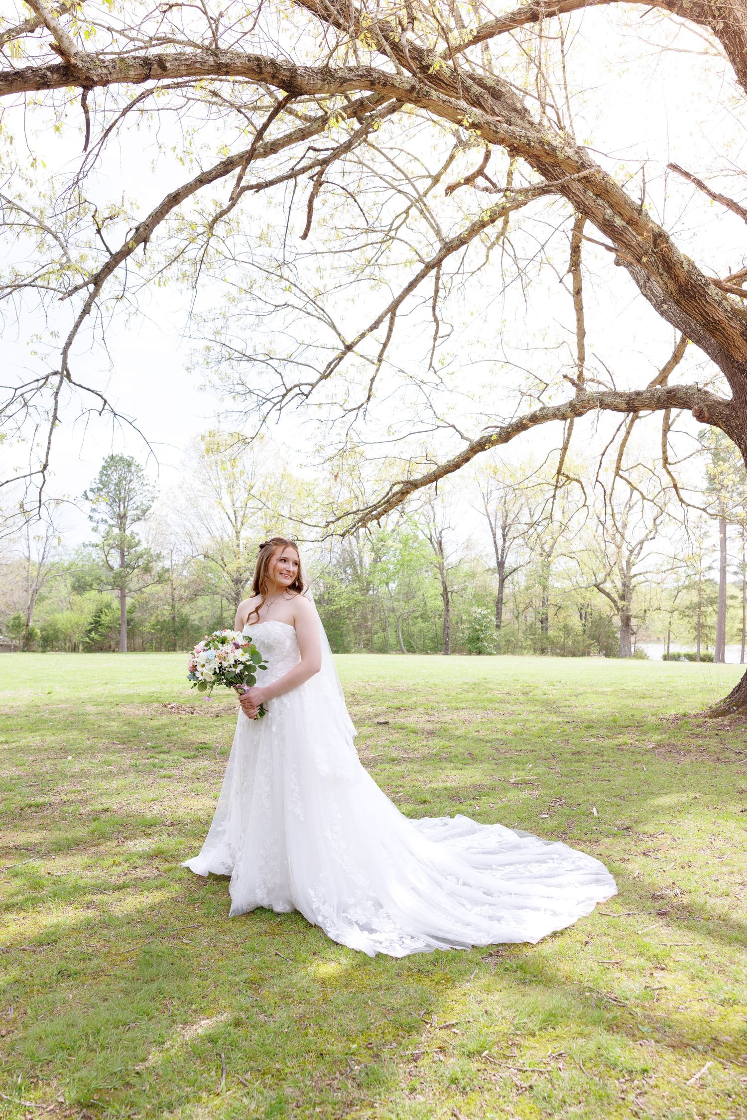 Bride in a white lace gown with a long train holds a bouquet under a large tree in a green outdoor setting.