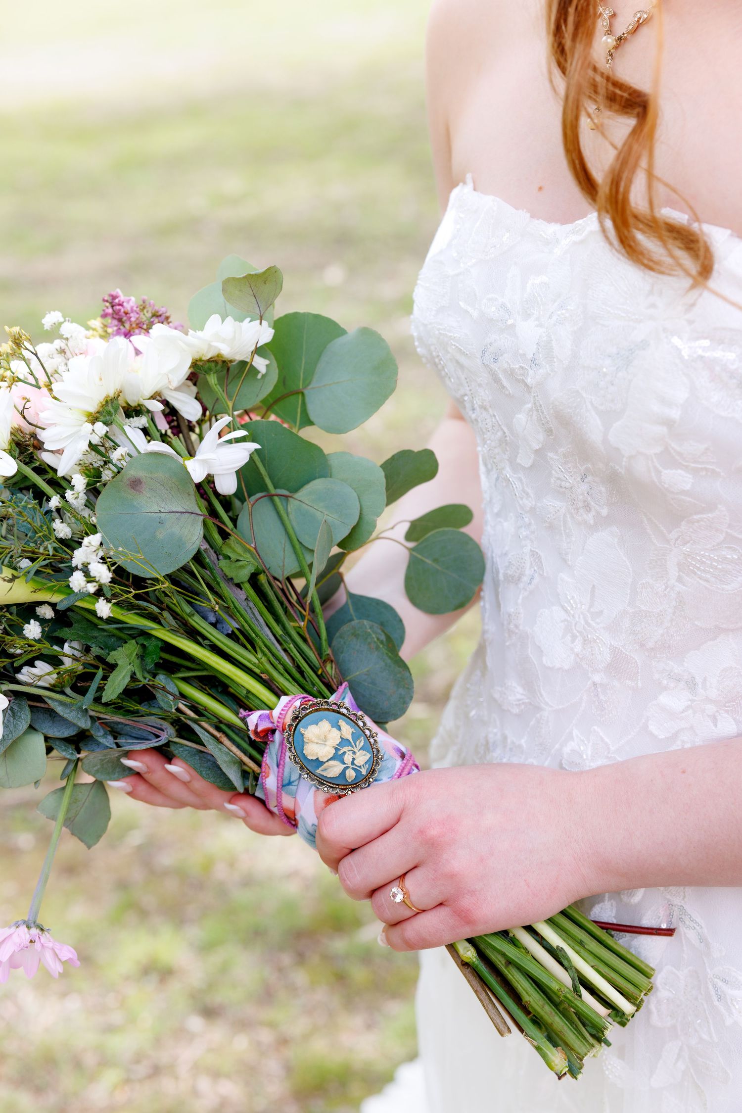 Bride in lace gown holding wedding bouquet of white flowers and eucalyptus with decorative brooch wrap.