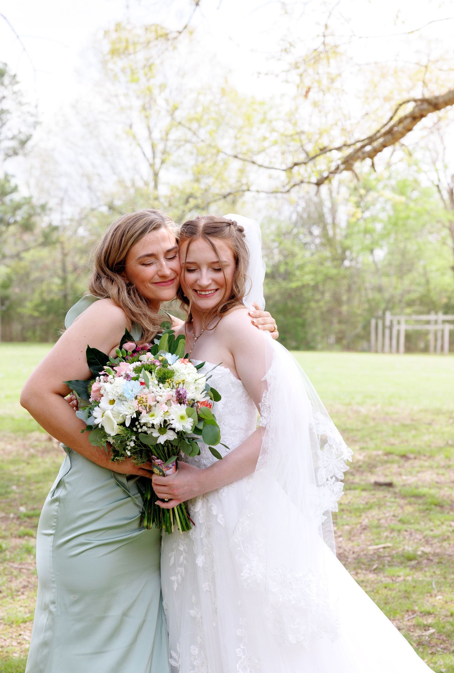 Bride in white lace wedding gown embraces bridesmaid in sage green dress while holding a floral bouquet outdoors.