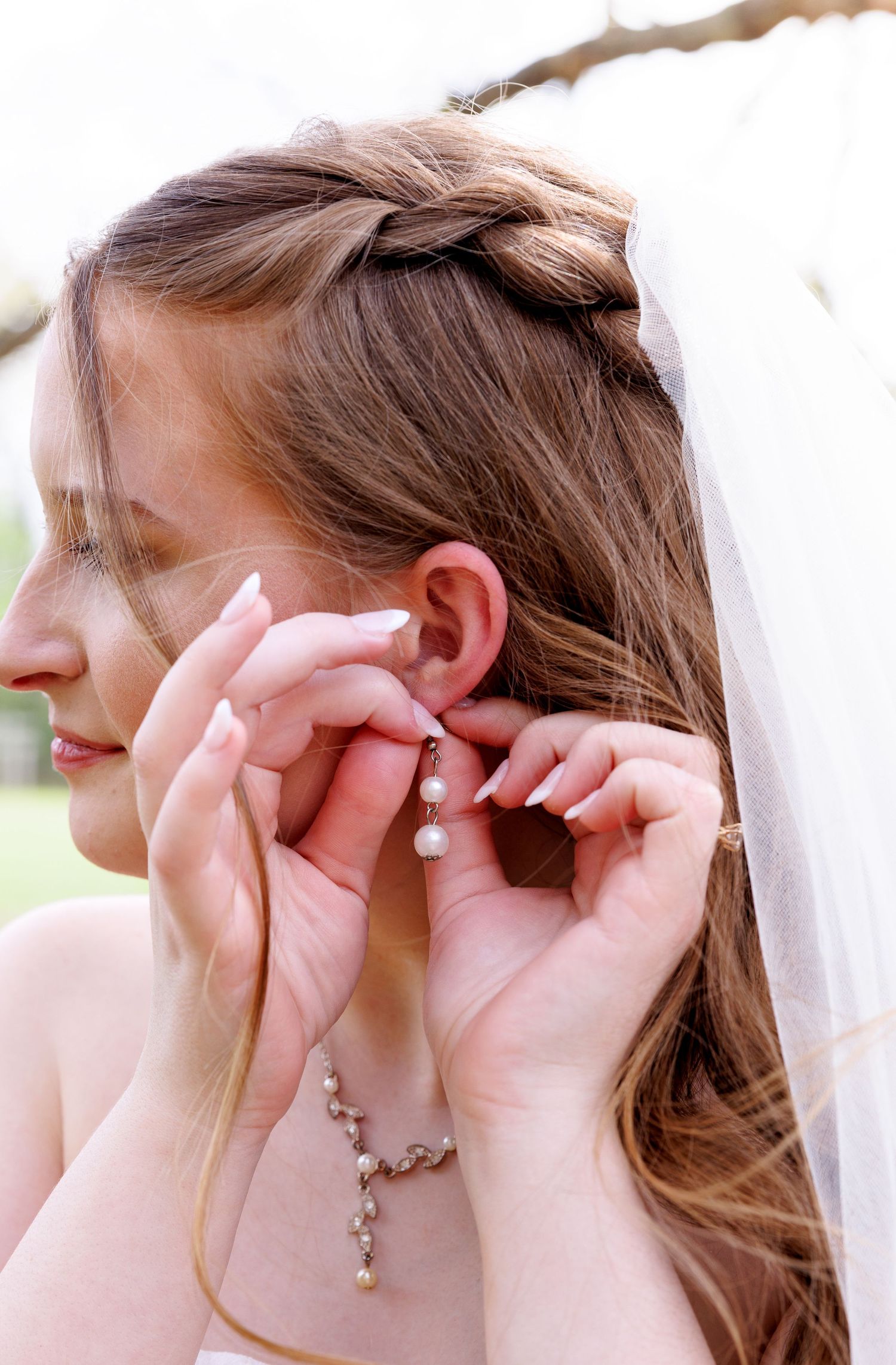 Bride with braided hair and veil putting on pearl drop earrings on her wedding day.