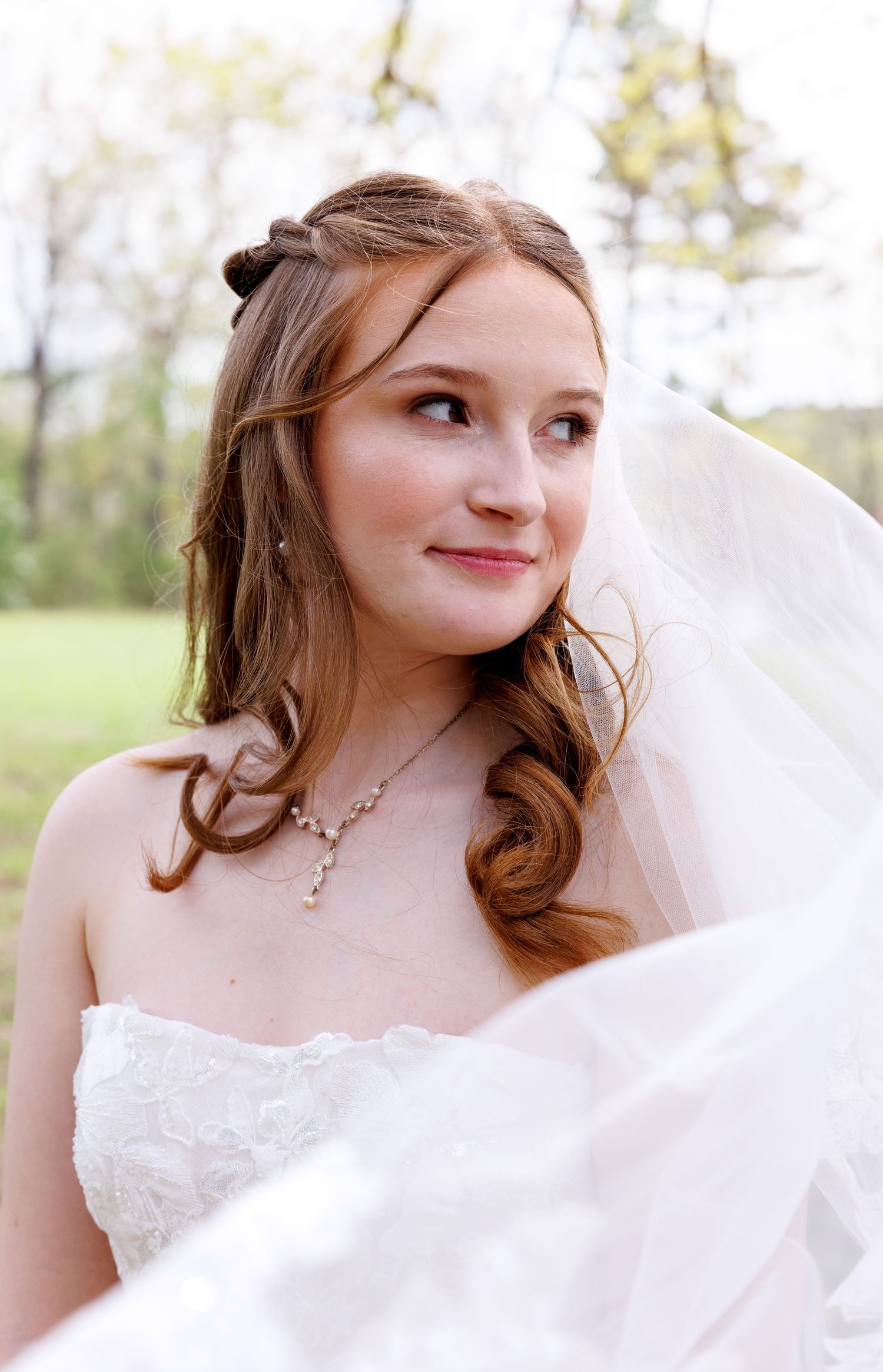 Young bride in white strapless gown and veil with pearl necklace smiling outdoors on her wedding day.