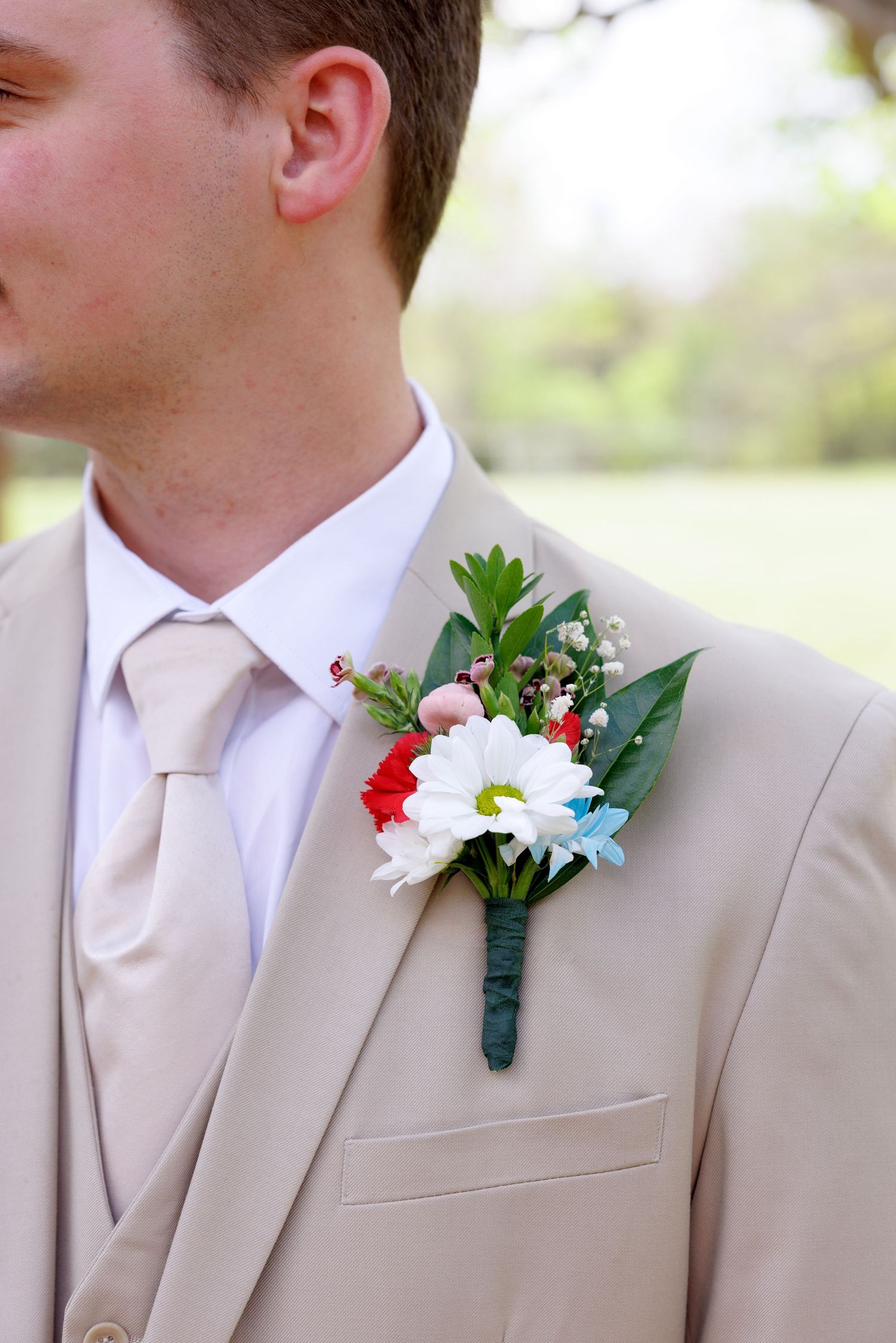 Groom in beige suit with colorful boutonniere featuring white daisy, red and blue flowers, and greenery on wedding day.