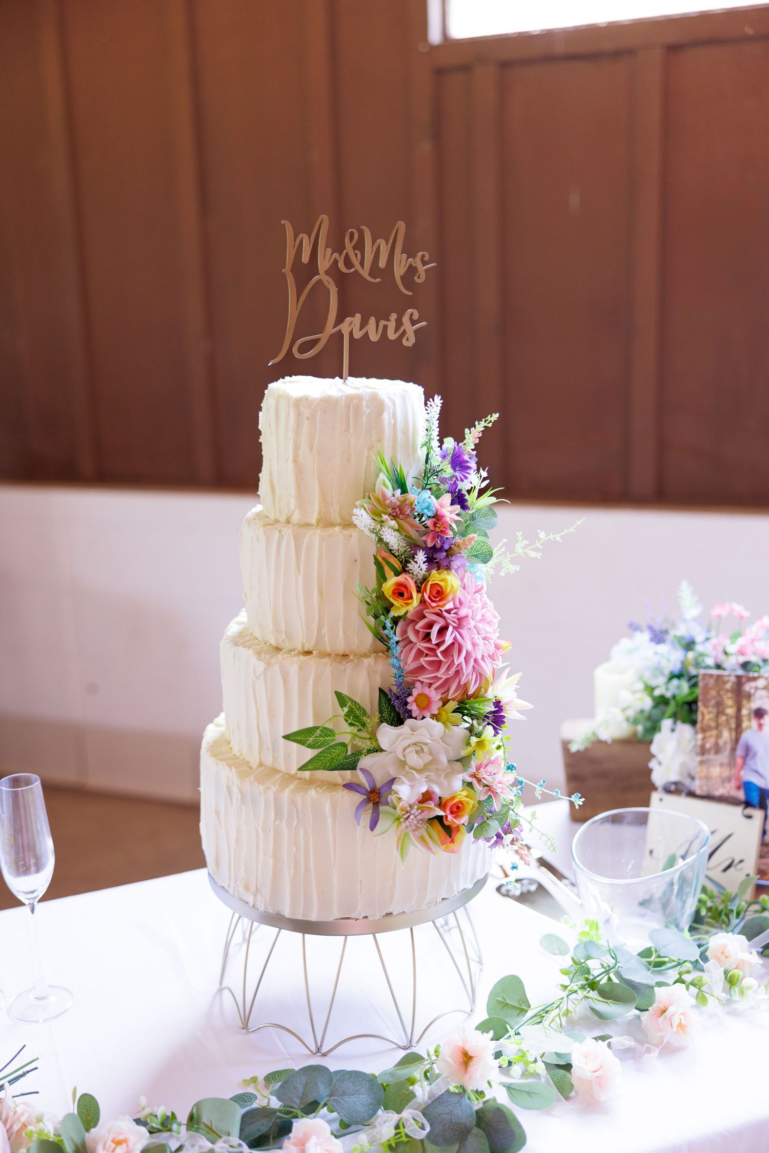 Four-tier white wedding cake with colorful floral decorations and a Mr. & Mrs. Davis gold cake topper on a table.