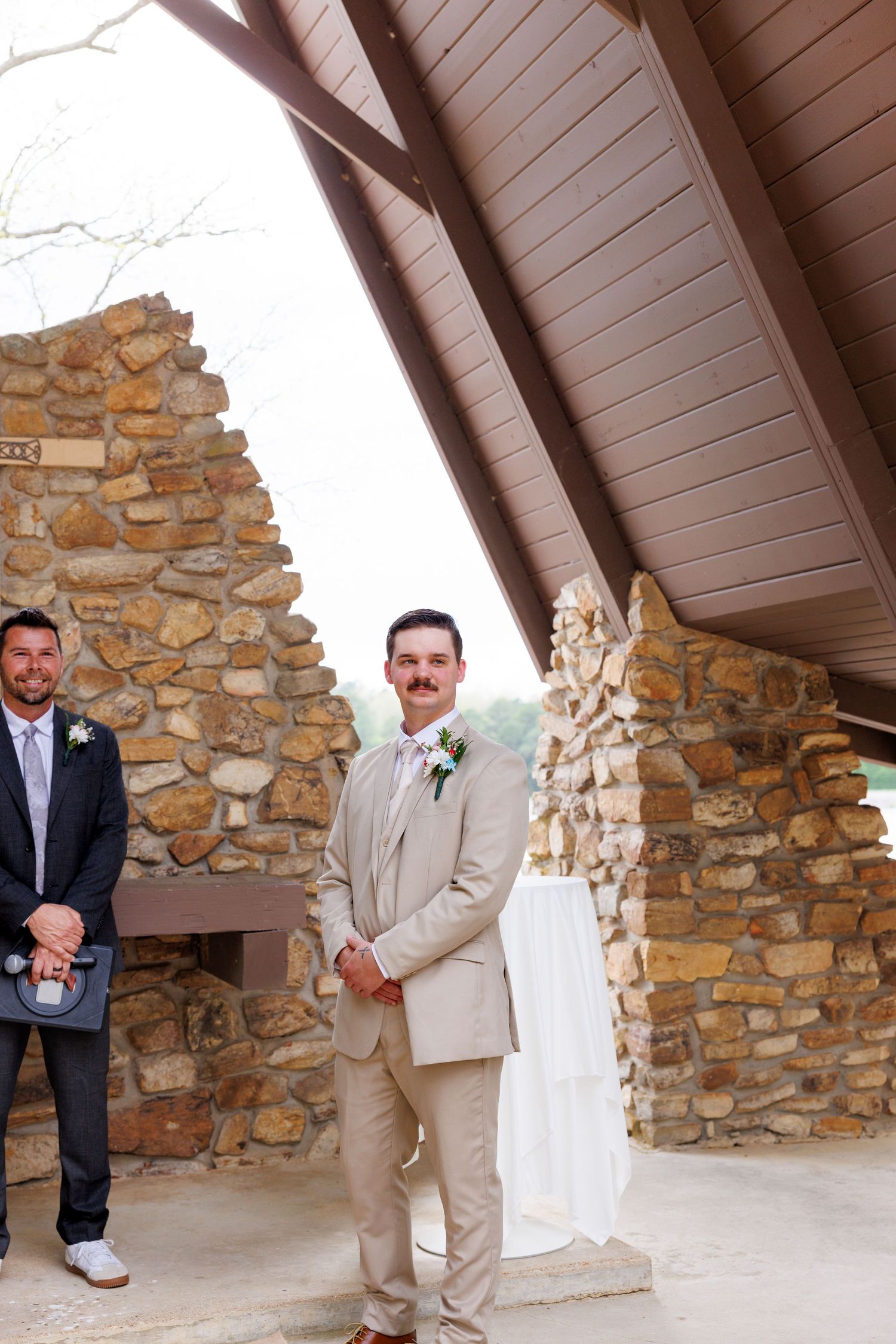 Groom in tan suit with boutonniere stands at outdoor stone chapel wedding venue with officiant nearby.