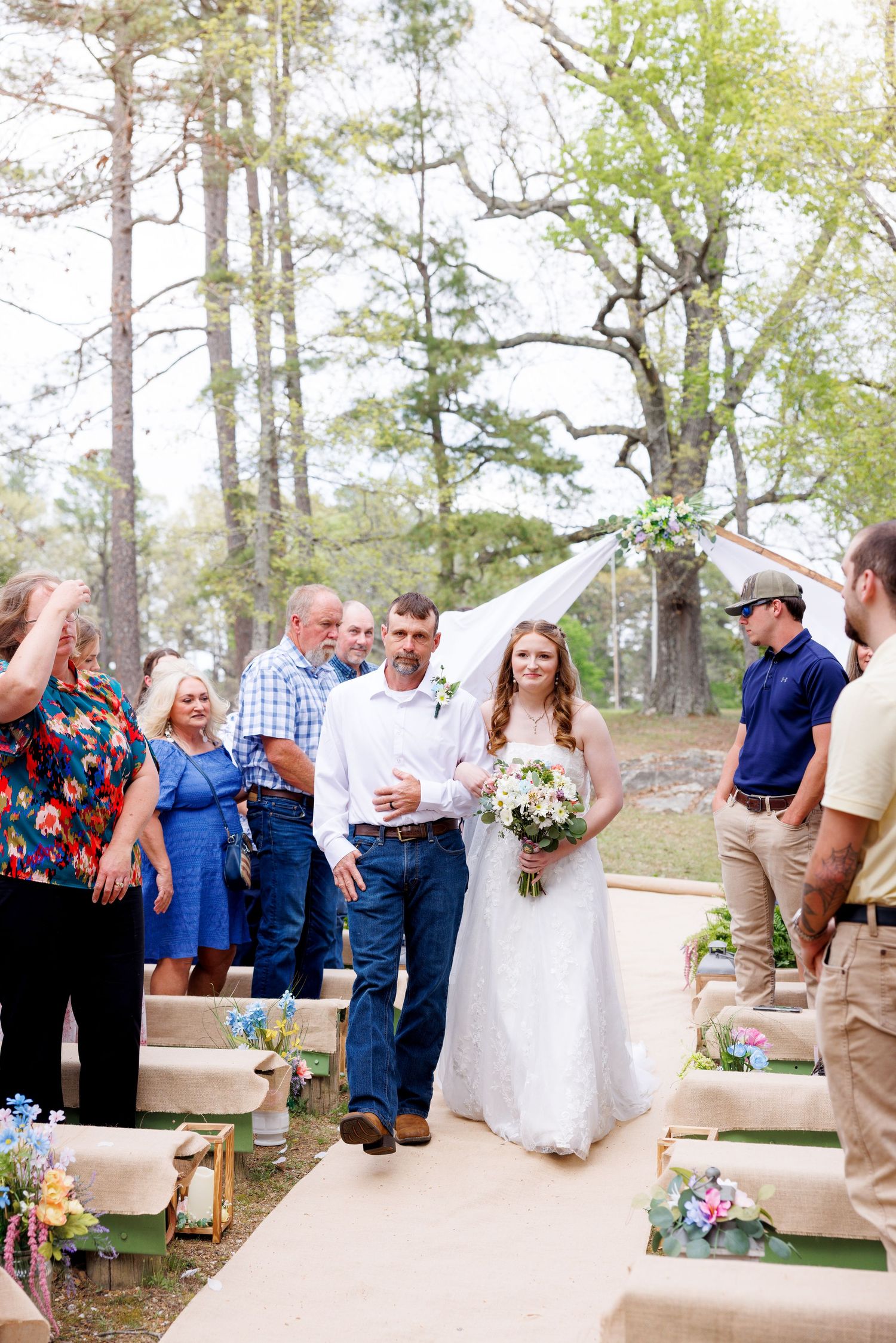 Bride in lace gown with bouquet walks down outdoor aisle with escort at rustic woodland wedding ceremony.