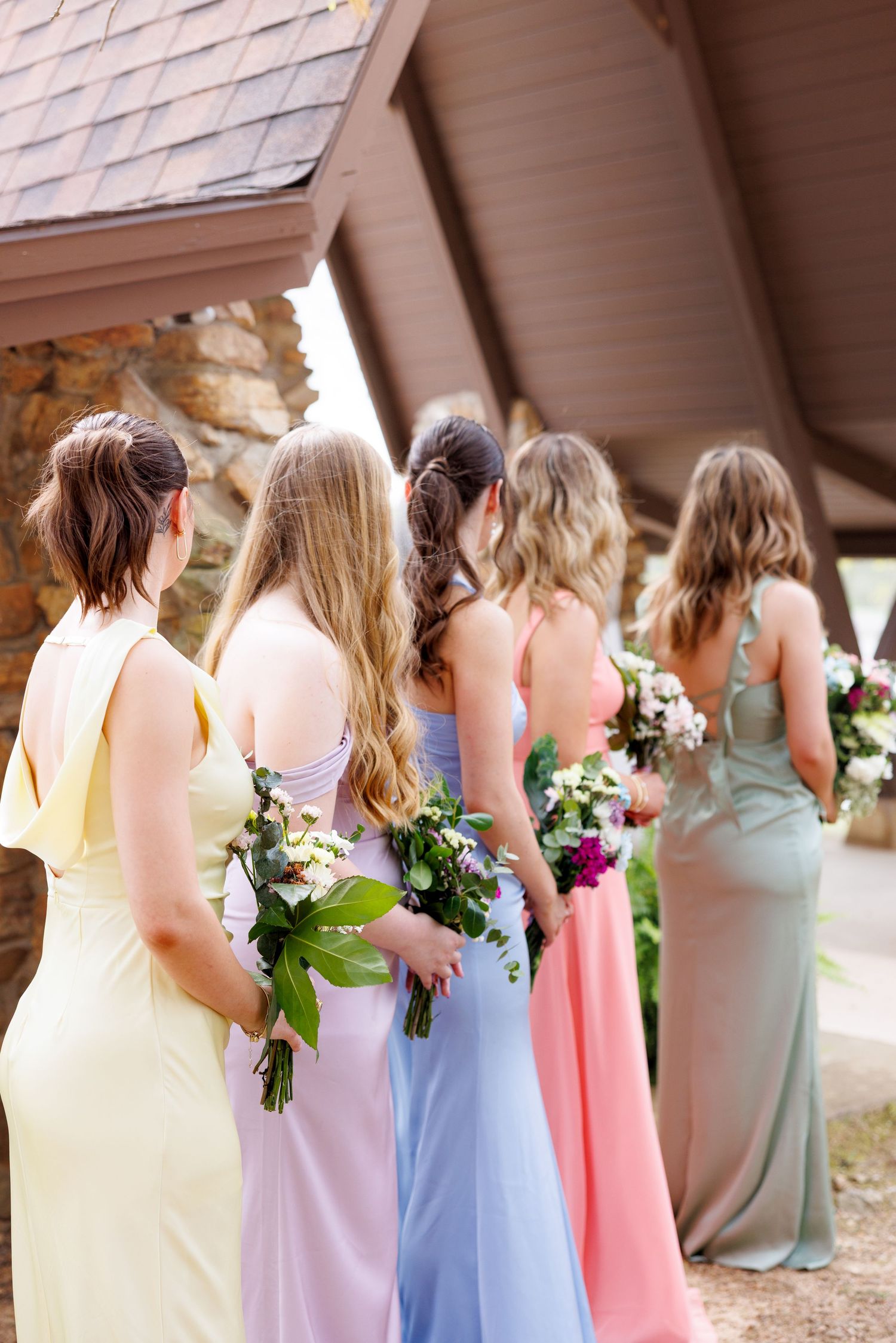 Bridesmaids in colorful mismatched pastel gowns holding bouquets stand with backs to camera outside rustic wedding venue.