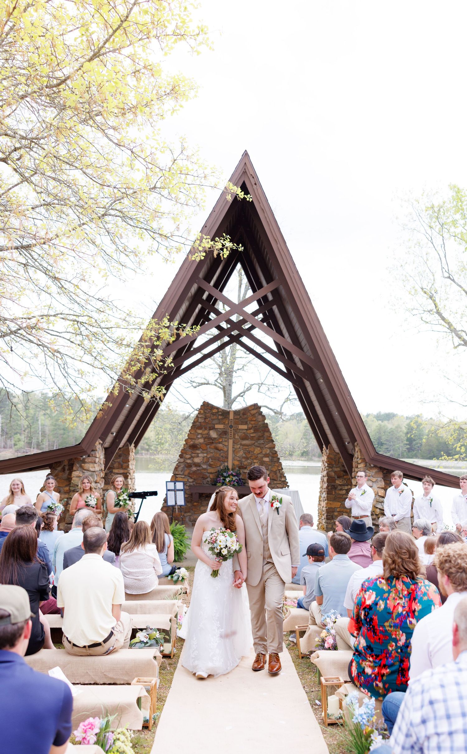Newlyweds walk down the aisle of an outdoor A-frame chapel by a lake surrounded by wedding guests.