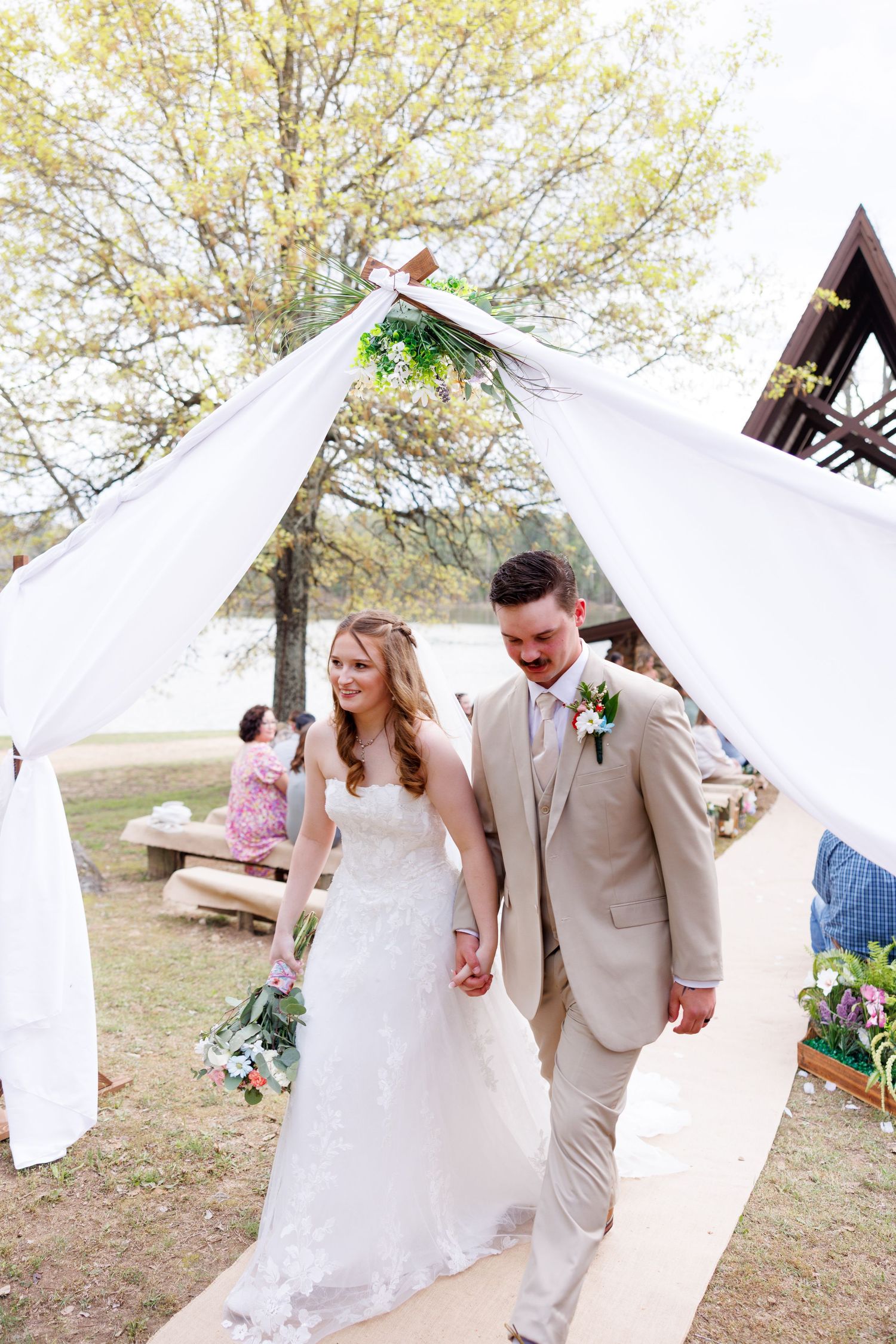Bride and groom walk hand in hand down the aisle under white draped fabric at an outdoor wedding ceremony.