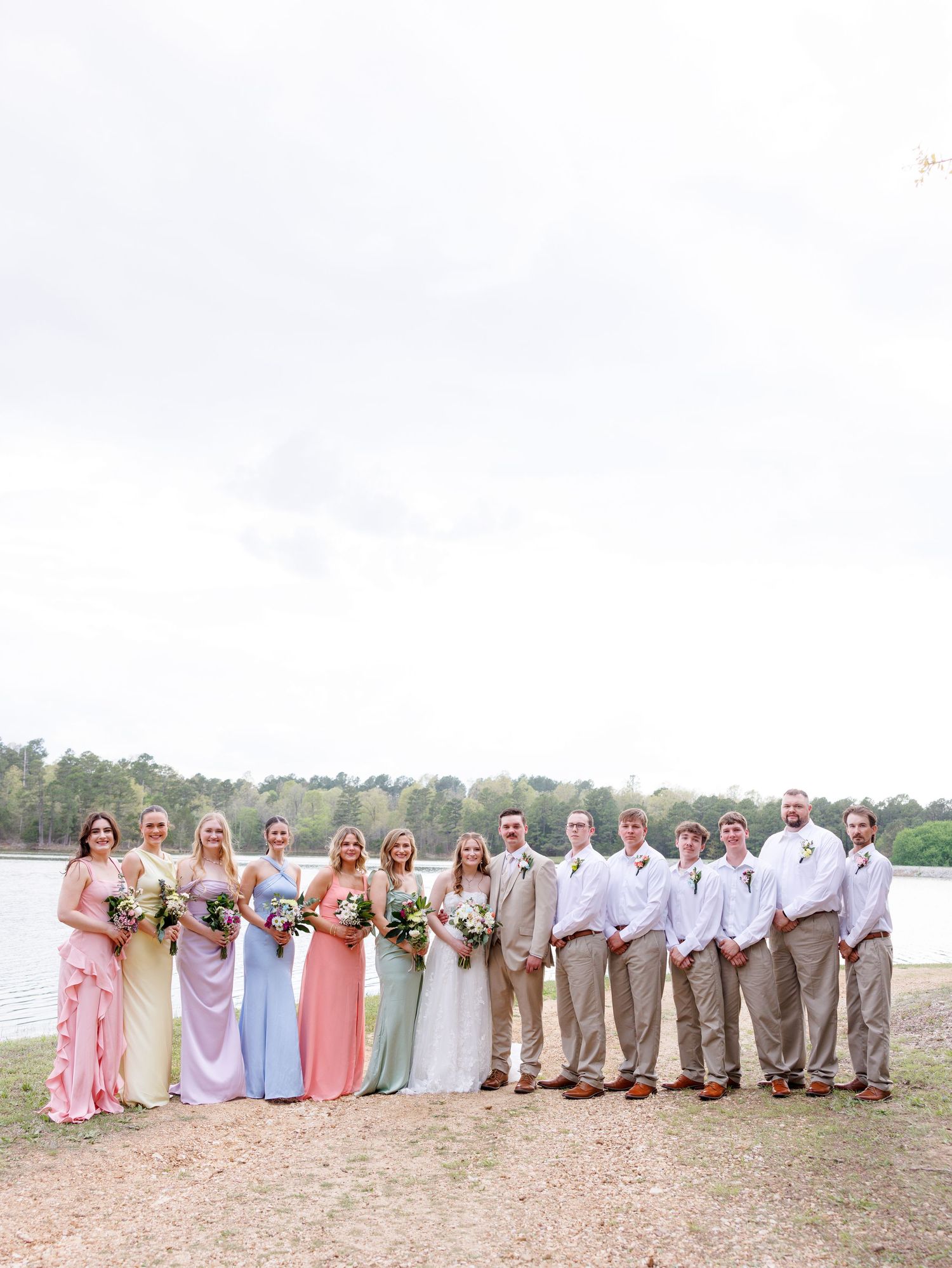 Wedding party poses by a lake, bridesmaids in pastel gowns and groomsmen in tan suits on a cloudy day.