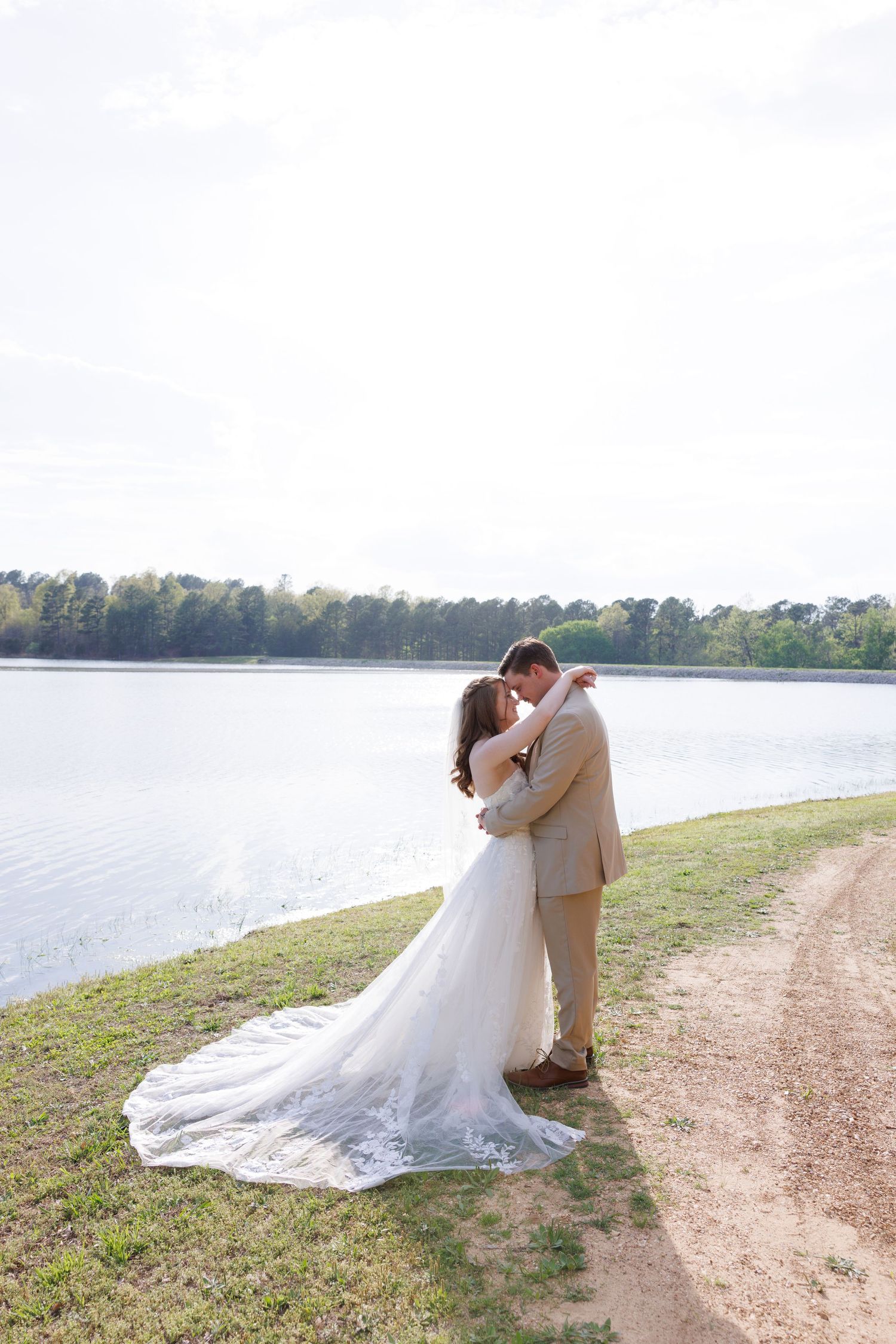 Bride and groom embracing by a serene lakeside, with her white lace wedding gown train spread across the grass.