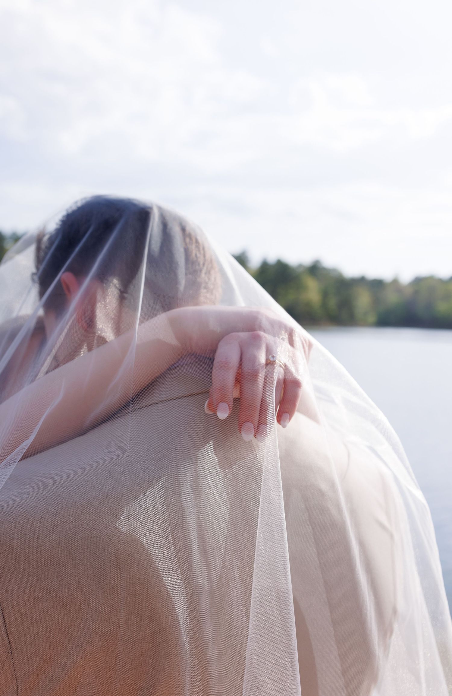 Bride in white veil gazing over a serene lake, her hand resting on her shoulder with a delicate ring.