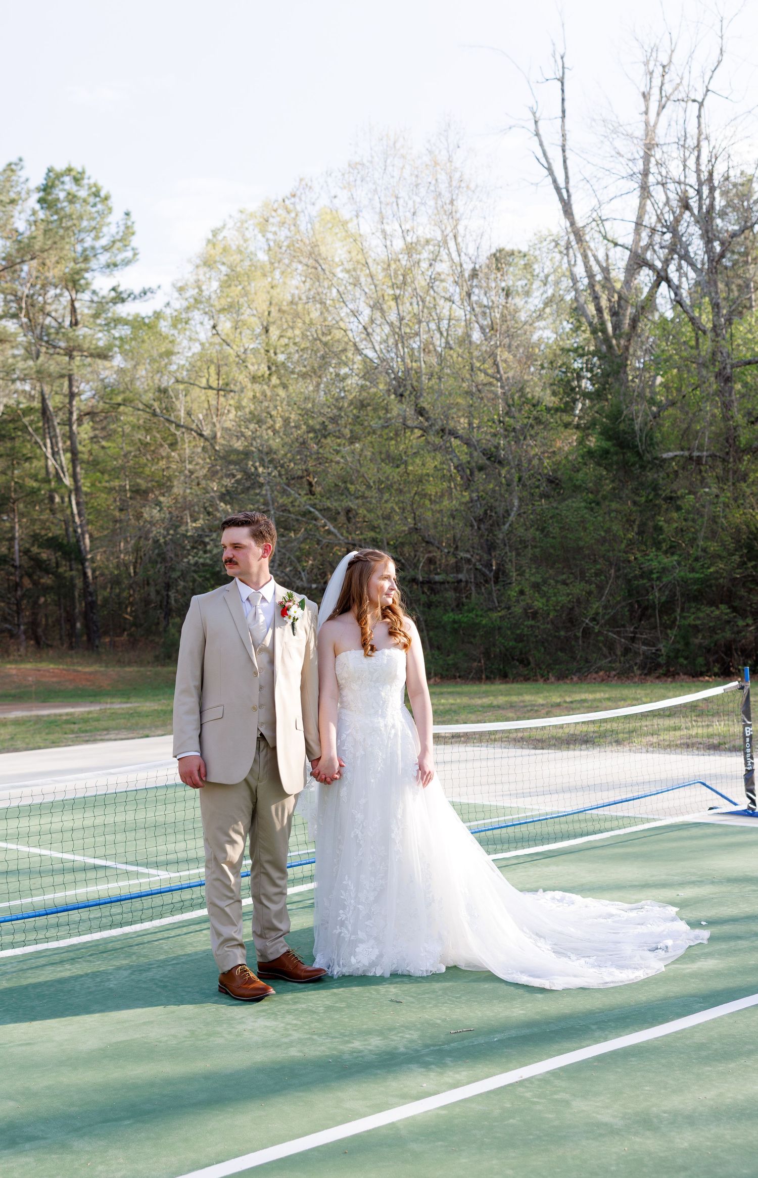 Bride and groom hold hands on a tennis court surrounded by trees in a romantic outdoor wedding portrait.