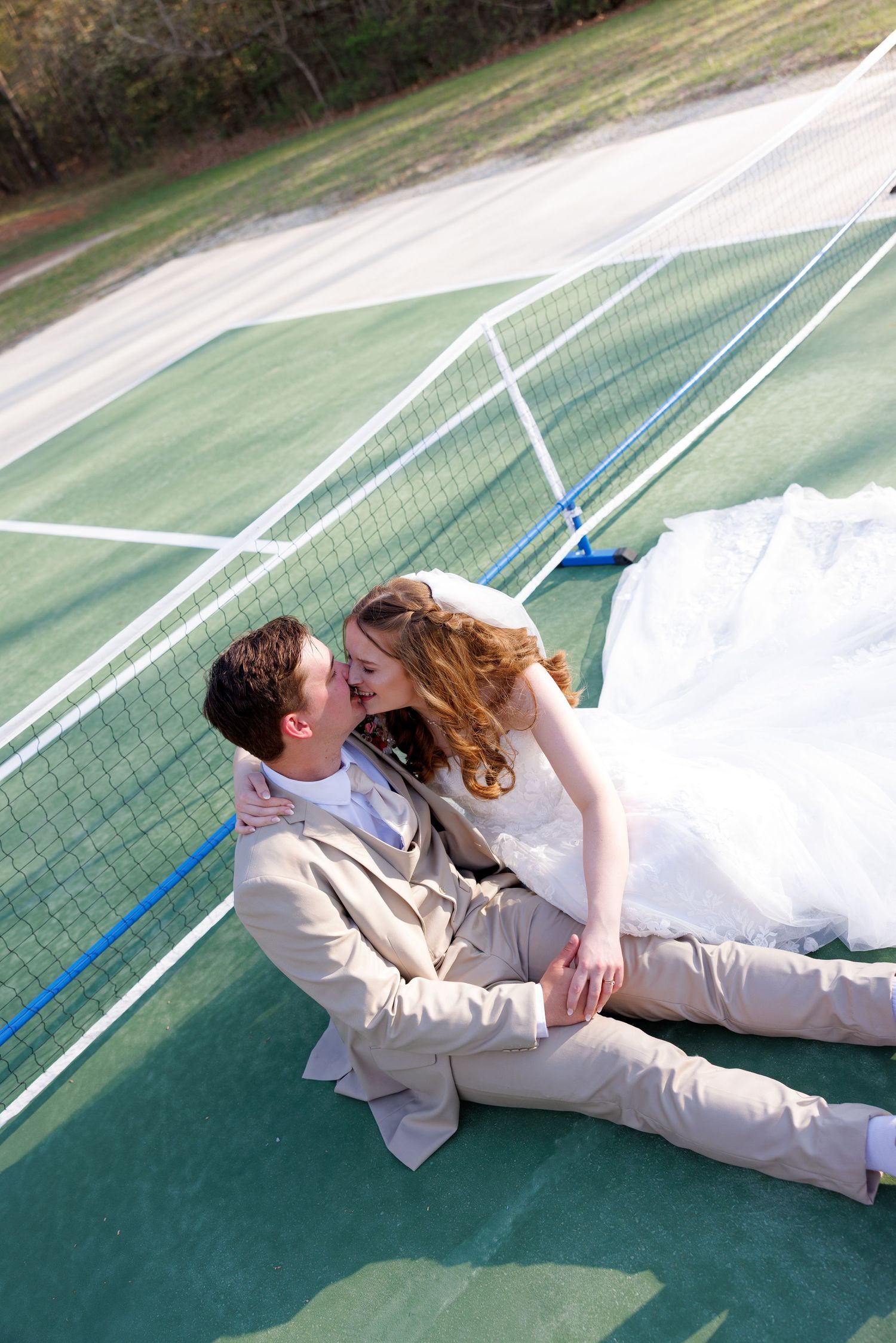 Bride and groom in wedding attire share a kiss while sitting on a green tennis court outdoors.