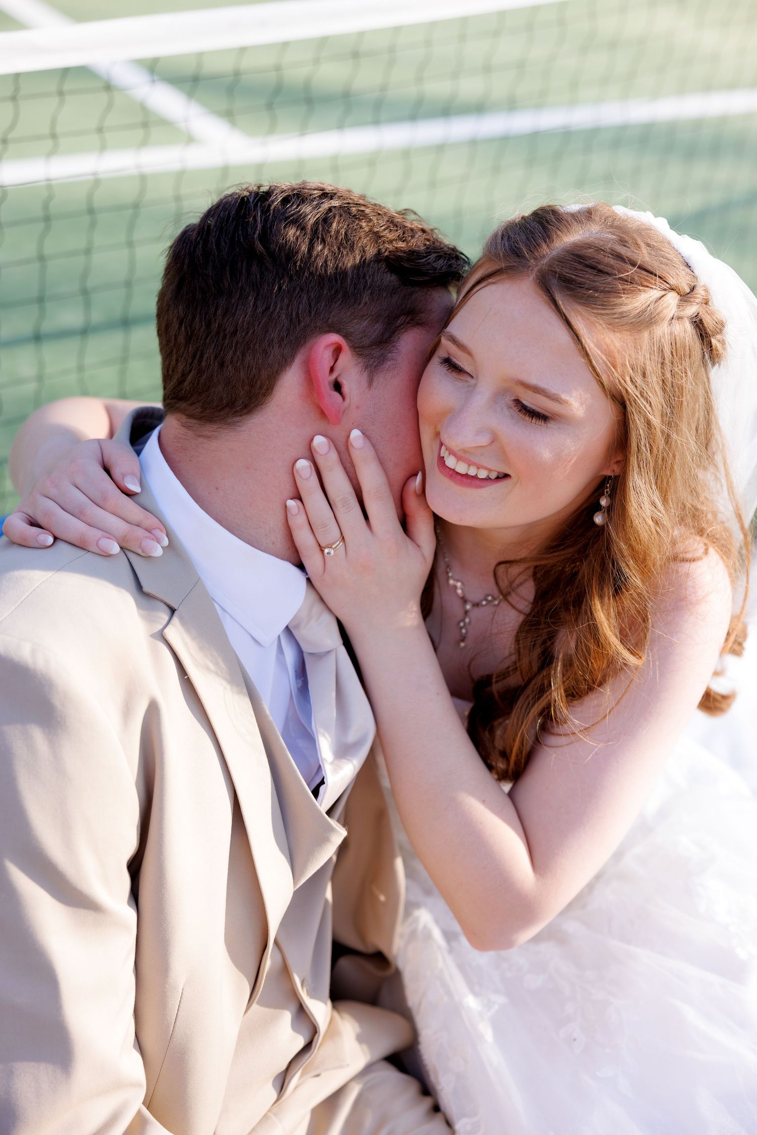 Smiling bride in white gown embraces groom in tan suit near a tennis court net in a romantic moment.