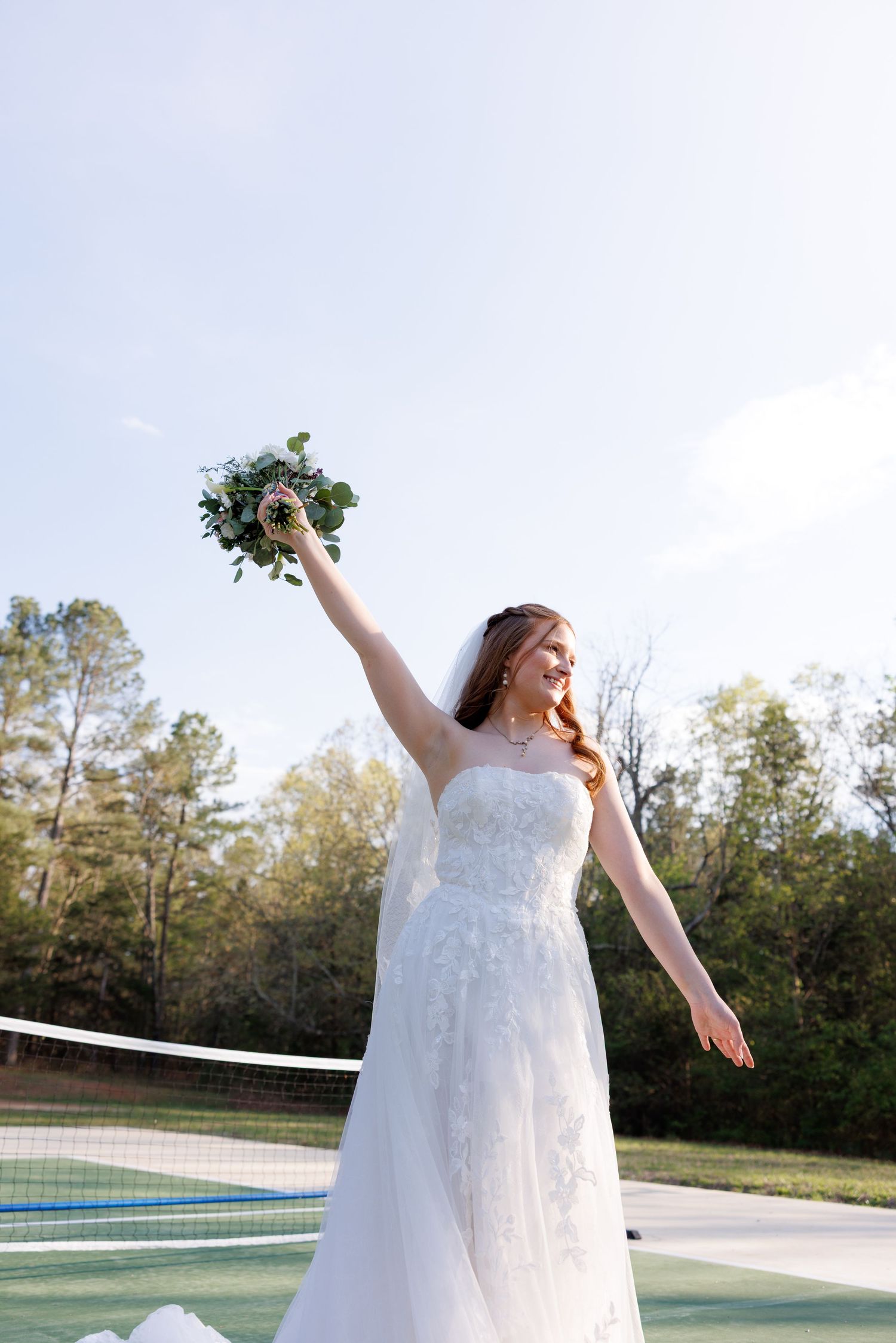 Joyful bride in white lace gown raises bouquet triumphantly on a tennis court with trees in the background.