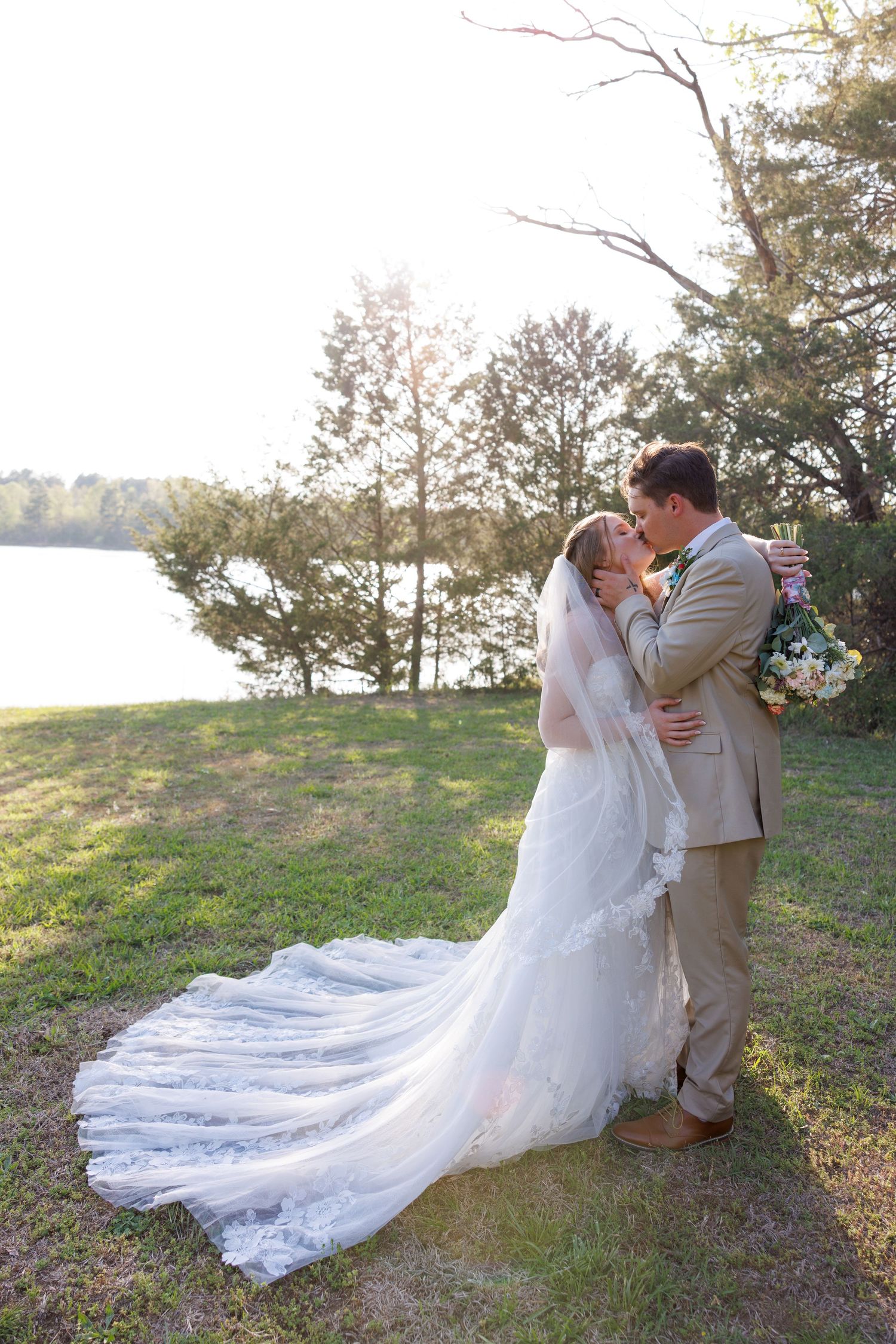 Bride and groom share a romantic moment by a lakeside, her lace train flowing across the grass at sunset.