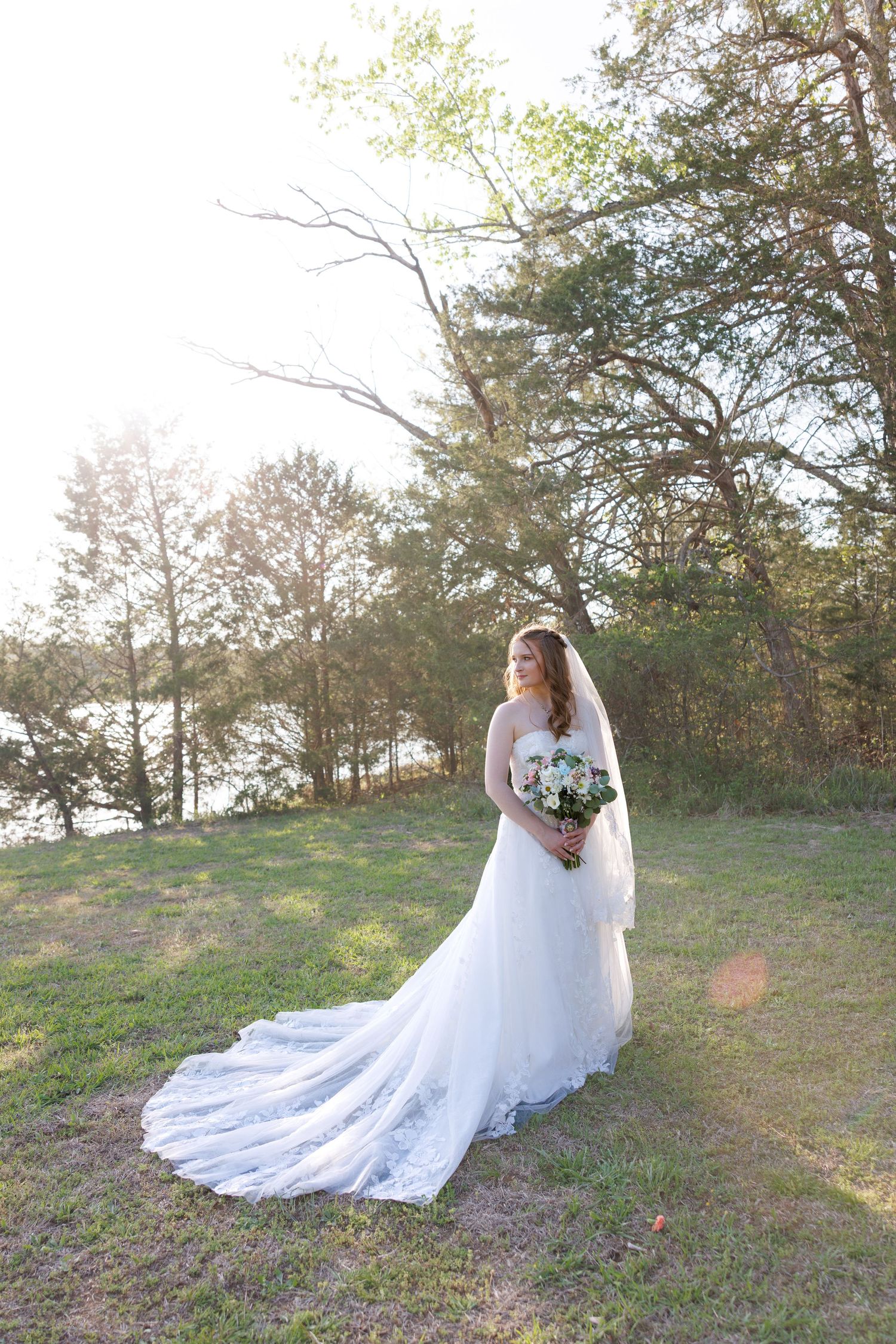 Bride in white lace gown with long train holds bouquet outdoors near lake and trees at golden hour.