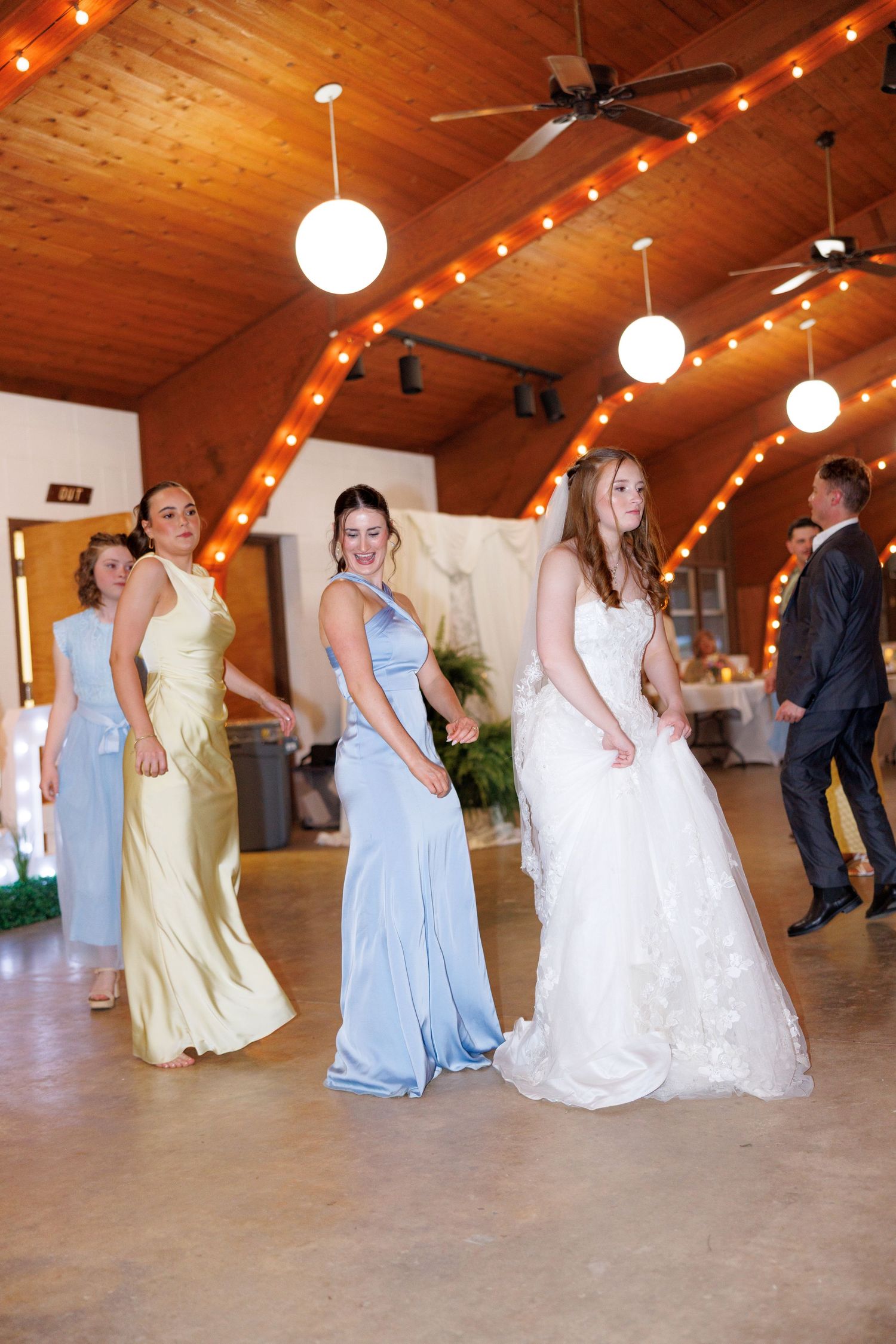 Bride in white lace gown dancing with bridesmaids in pastel dresses at a rustic wedding reception.