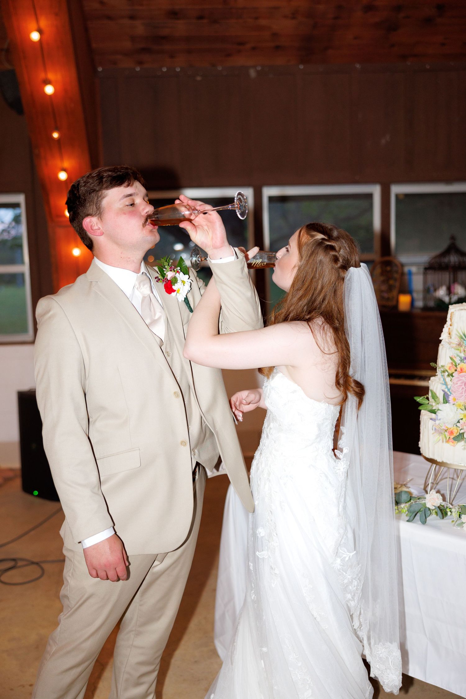 Bride and groom share a playful champagne toast near wedding cake at rustic barn reception.