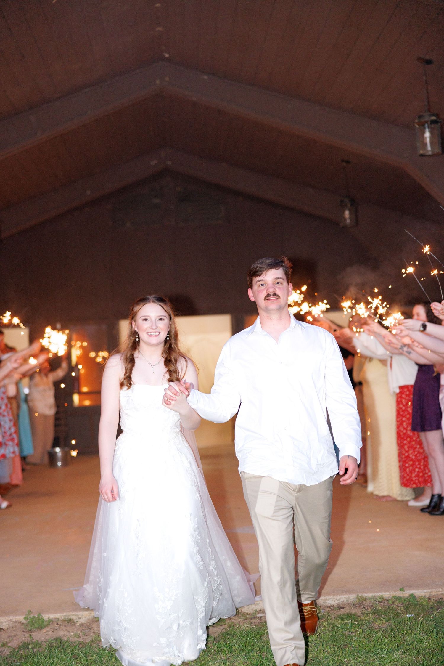 Newlyweds walk through sparkler send-off tunnel outside rustic barn wedding venue at night.