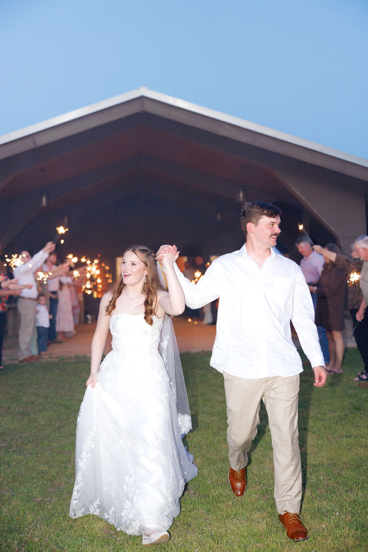 Newlyweds holding hands walking through a sparkler send-off exit outside a wedding reception venue at dusk.