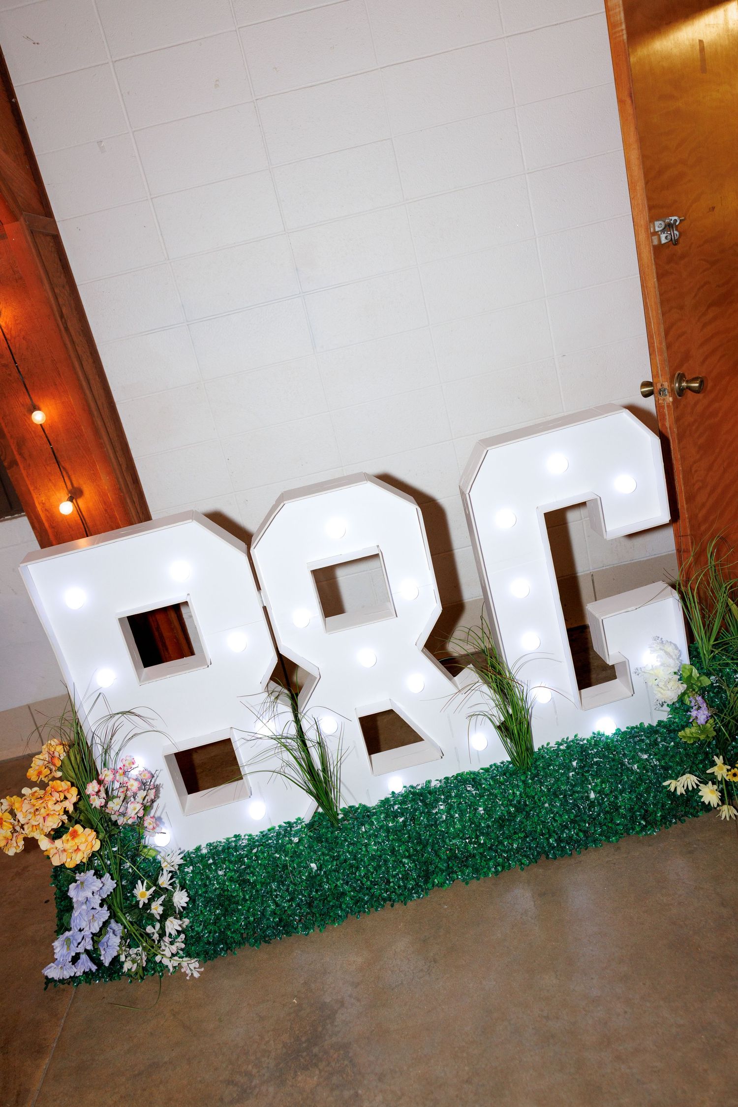 Large illuminated marquee letters B&C with floral decorations on grass display at wedding venue.