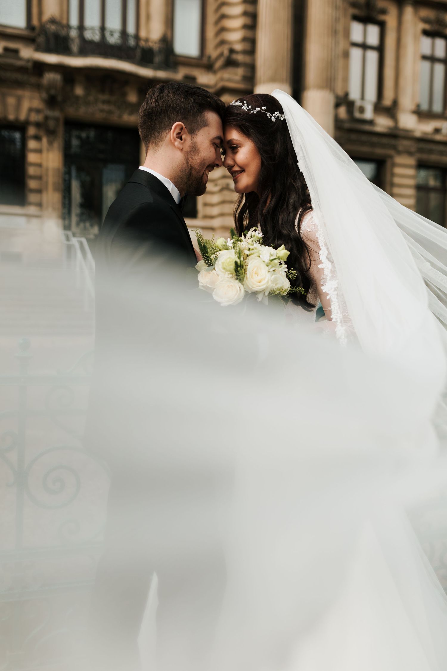 Bride and groom share a romantic forehead touch moment, veil flowing, holding white bouquet outdoors.