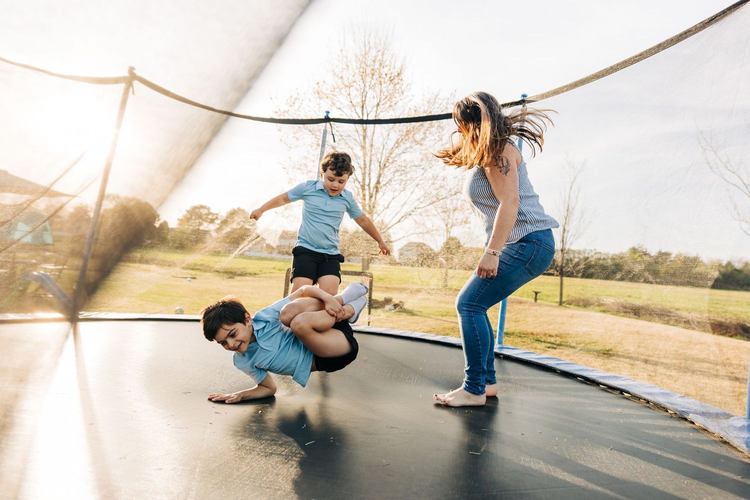 Two kids playing and jumping on a large outdoor trampoline with safety net on a sunny day with their mom in Charlotte, NC.