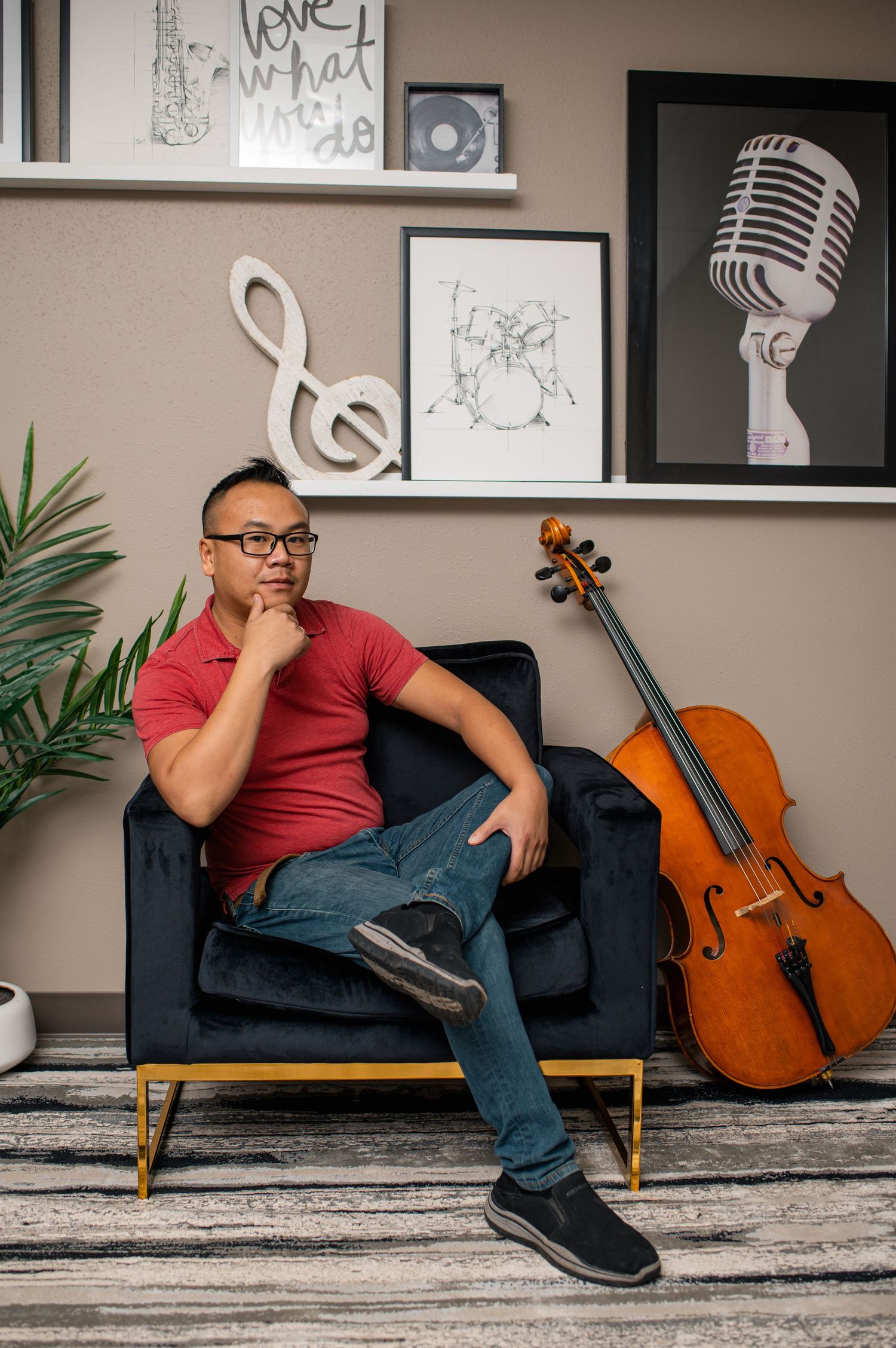 Man in red shirt sits thoughtfully in velvet chair beside a cello, surrounded by music-themed wall decor.