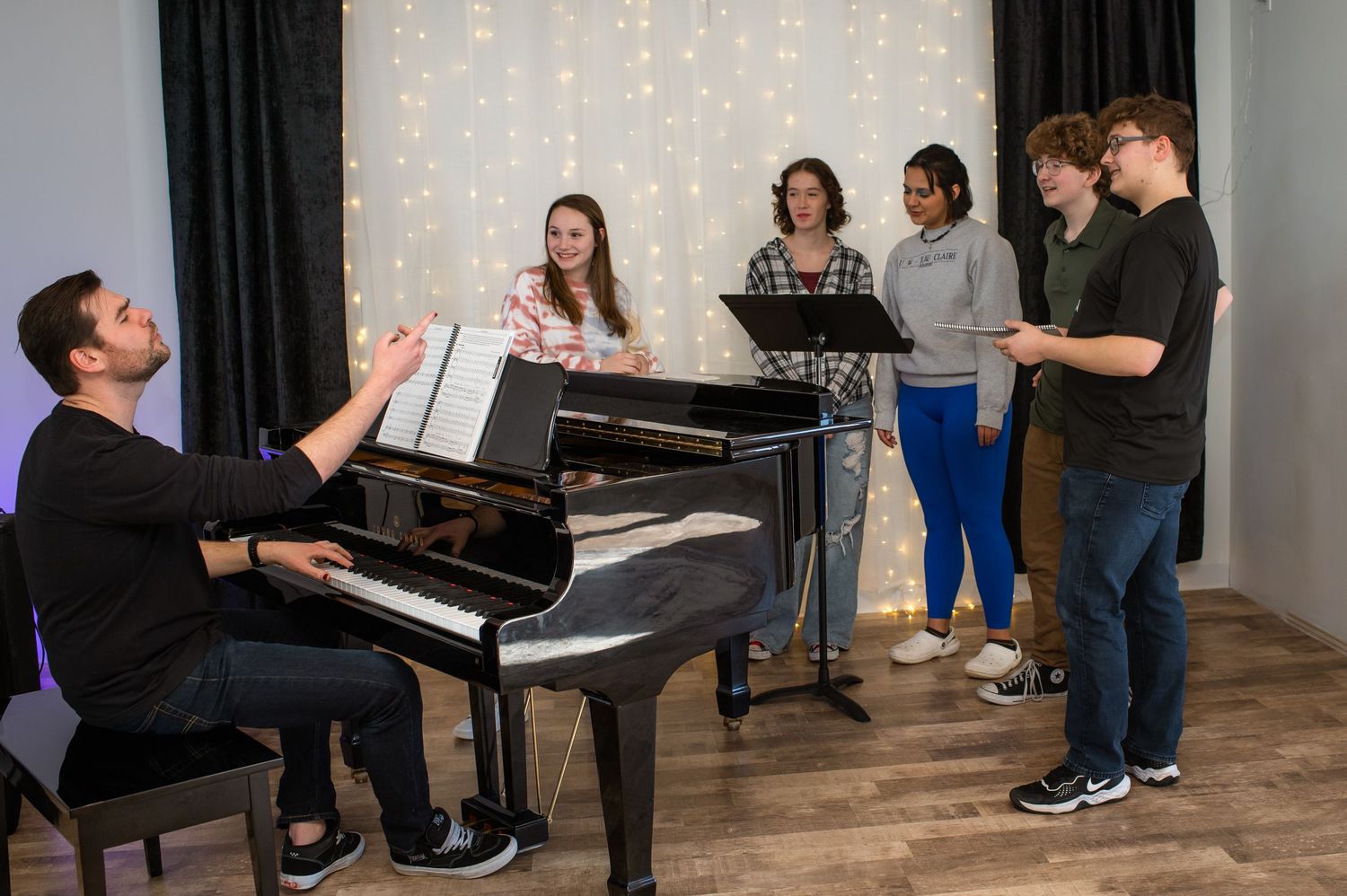 Students gather around a grand piano during a vocal music rehearsal in a warmly lit studio with fairy lights.